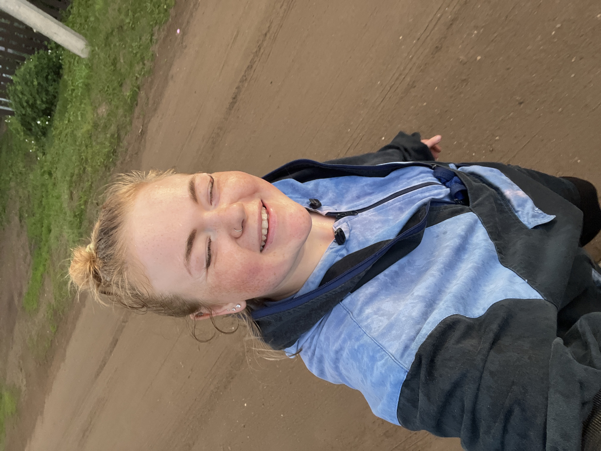The image is a close-up, outdoor, selfie-style shot of a young woman with blonde hair pulled back in a bun. She is wearing a light blue and black jacket. Her eyes are closed, and she has a wide smile, suggesting happiness or amusement. Freckles are visible across her nose and cheeks, and she has a nose ring and multiple earrings. The background appears to be a dirt path or road, with some green grass and a fence visible on the left side. The lighting suggests it might be daytime, possibly overcast given the absence of strong shadows. The image is oriented vertically, with the woman's head at the bottom of the frame. There is no visible text in the image. The location is specified as Olkhovka, Russia.