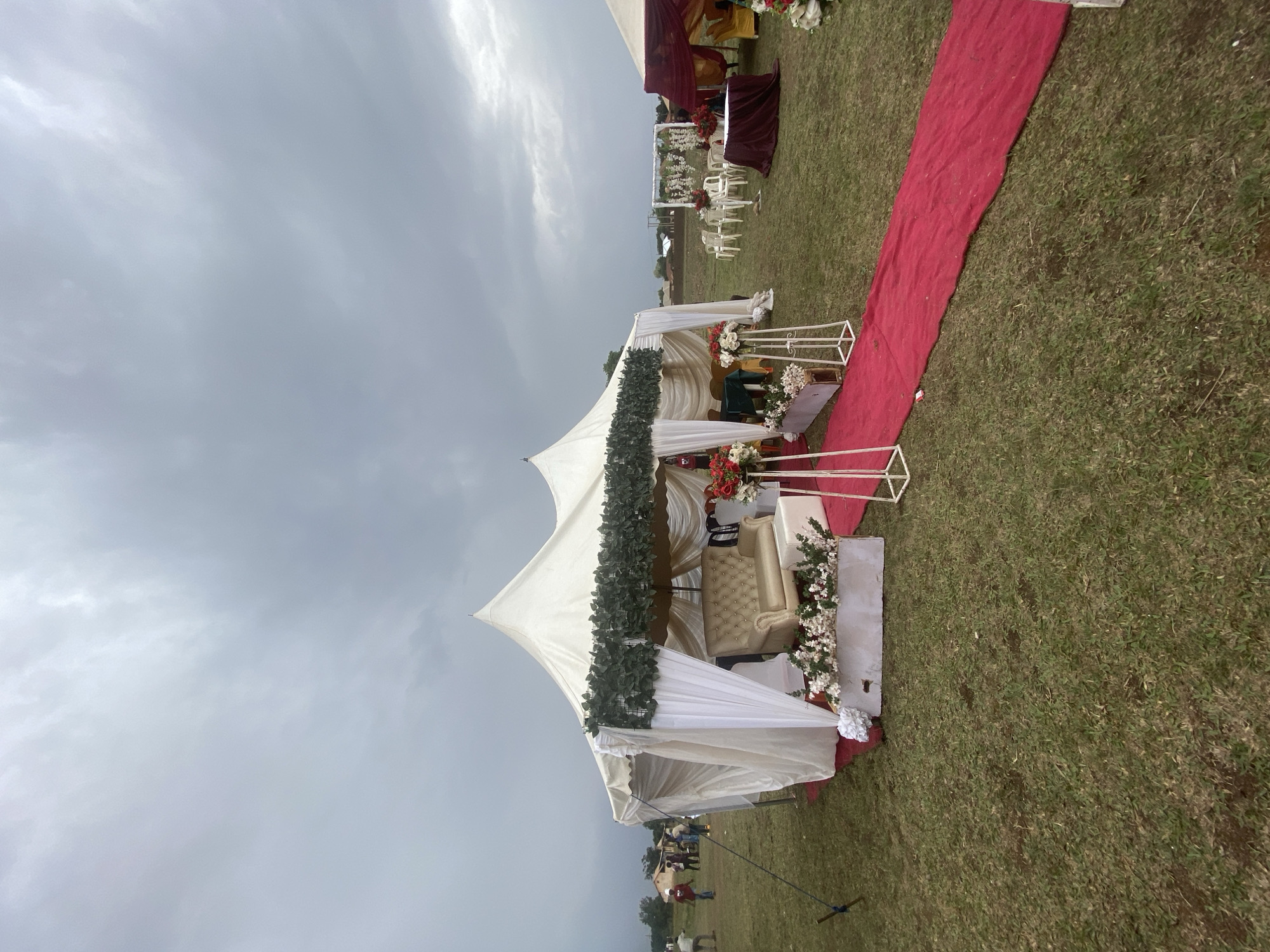 The image depicts an outdoor event, possibly a wedding, in Kuru Dungus, Nigeria. A white tent structure, decorated with greenery and white fabric, stands on a grassy lawn. A red carpet leads to the tent. A seating arrangement is visible inside the tent. In the background, more white chairs and tables are visible. The sky is overcast, suggesting the possibility of rain. Several people are standing in the background.