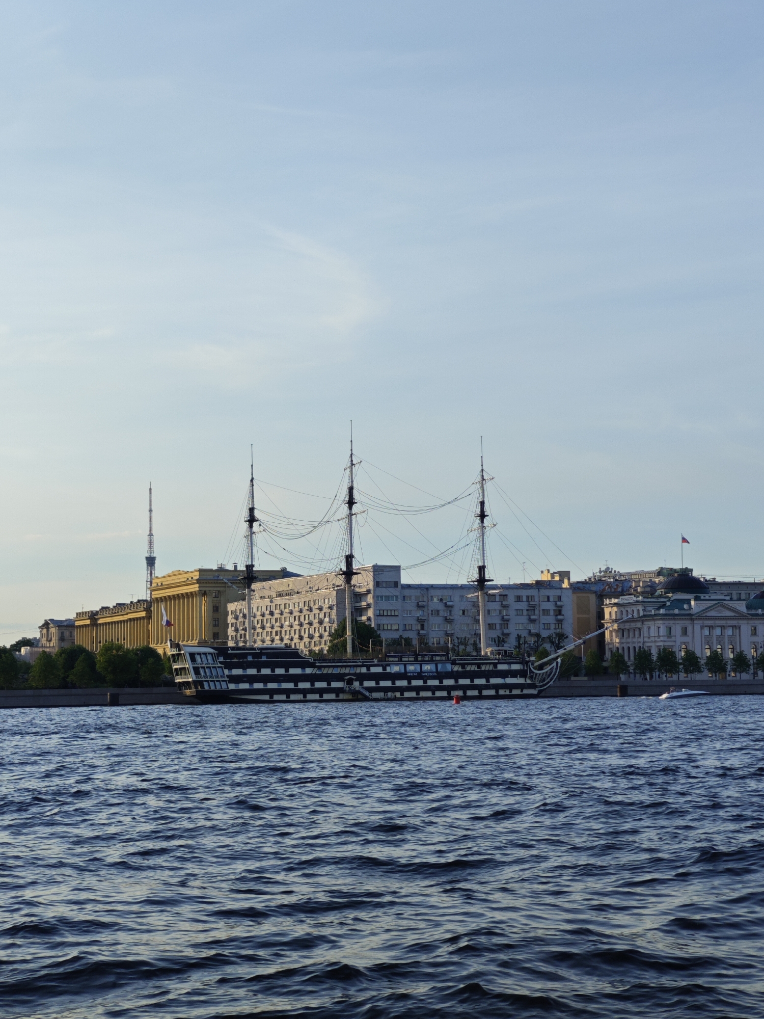 This is a photograph taken in Saint Petersburg, Russia, likely from a boat on the Neva River. The scene is dominated by a large, dark-hulled ship with multiple masts and rigging, docked near the waterfront. Behind the ship, a cityscape unfolds, featuring a mix of architectural styles. Prominent buildings include a structure with a classical colonnade, another with a modern facade, and a distant television tower. The sky is a pale blue, with some wispy clouds. The water in the foreground is dark blue and rippled, suggesting gentle waves. The overall impression is one of a historical naval vessel set against the backdrop of a modern urban environment.