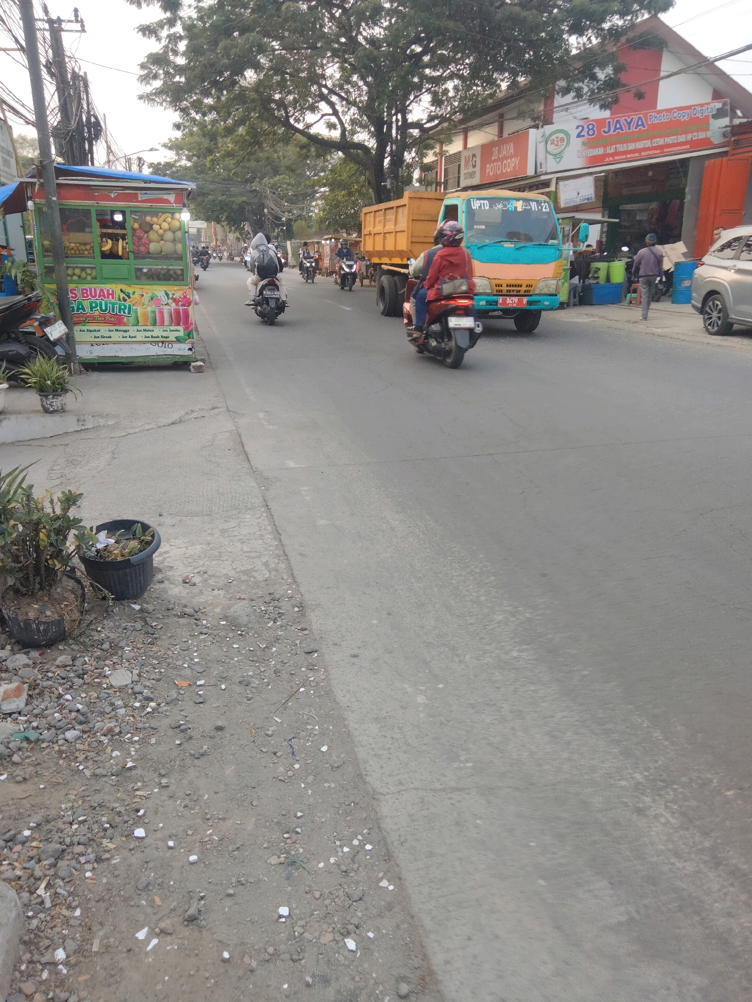 The image depicts a street scene in Sepatan, Indonesia. In the foreground, on the left side of the road, is a fruit juice stand labeled "BUAH SA PUTRI". The stand is colorful and displays various fruits, including mangoes, bananas, and guavas. Adjacent to the stand are potted plants and a pile of gravel.
The road itself is paved and shows signs of wear and tear. Traffic is present, with several motorcycles and a large truck visible. A motorcycle with two riders is in the immediate foreground, moving away from the camera. Another motorcycle is parked near the fruit stand. In the middle ground, a blue and orange truck is driving on the road. In the background, buildings line the street, including a building with signage that reads "28 JAYA Photo Copy Digital". The environment is illuminated by natural daylight, suggesting it is daytime. The overall impression is of a busy, everyday street in a commercial area of Sepatan.