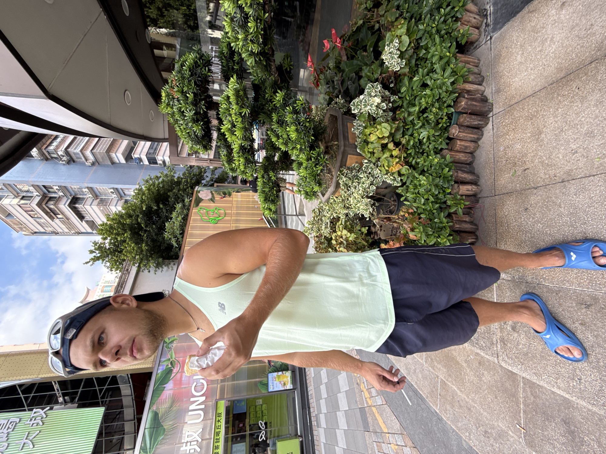 A young Caucasian male stands on a tiled outdoor walkway, eating an ice cream cone, in front of a building with a curved facade and a section of lush greenery. He is wearing a light green tank top, navy blue shorts, a black baseball cap with goggles resting on top, and blue flip-flops. The male is holding the ice cream cone in his right hand, and a white cigarette in his left hand. In the background, a tall blue and white building with many windows and air conditioning units is visible, along with trees and the facade of the building he is standing in front of. The building facade features large glass windows with green signage, and a neon green outline of a frog and flowers. The greenery includes various potted plants, some with red flowers, and a bonsai tree. The walkway is made of light brown tiles, and a street runs alongside it. The scene appears to be in Haizhu District, China.