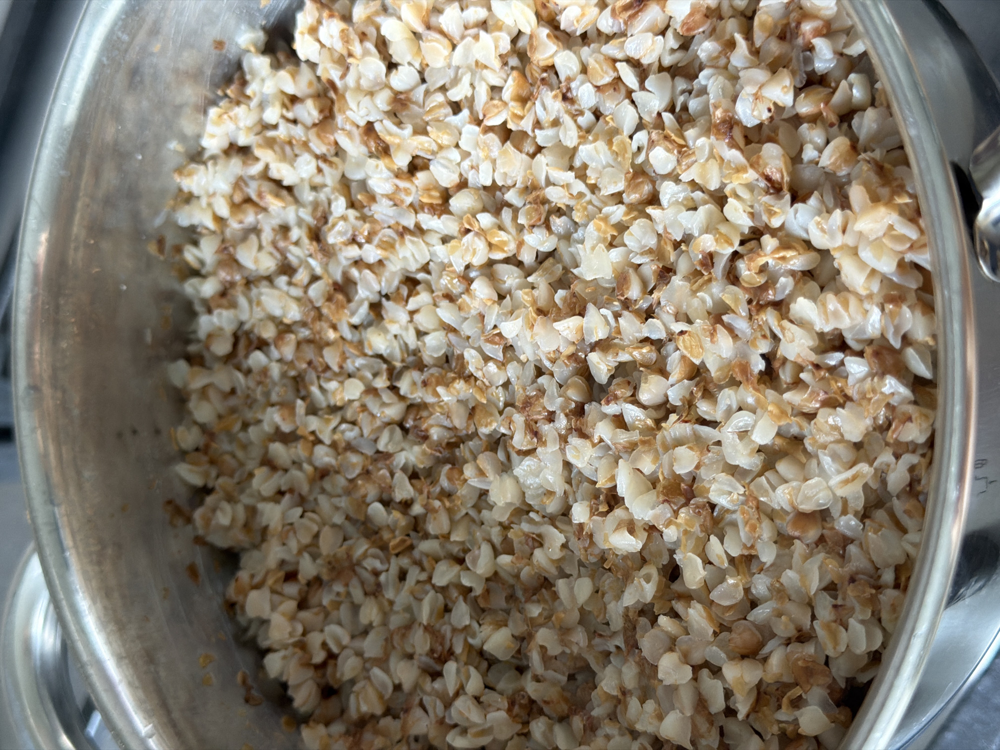 A top-down, close-up shot shows a pot filled with cooked buckwheat groats. The groats are a light brown and off-white color, with some appearing slightly darker and toasted. The pot is made of stainless steel with a visible rim. The background is out of focus, showing a hint of a stovetop. This image does not provide enough information to determine its location in Izhevsk, Russia.