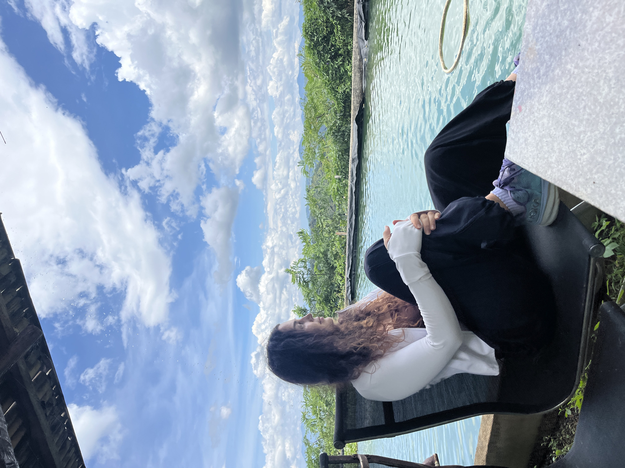 A woman with long, curly brown hair sits in a black reclining chair next to a pool of blue water. She is wearing a white long-sleeved shirt, a brown camisole, and black pants. Her legs are crossed, and she is looking off to the right. The pool is surrounded by lush green vegetation, and in the background, there are mountains under a bright blue sky with white clouds. The scene appears to be an outdoor relaxation area. The location is in Son Dien city, Vietnam.