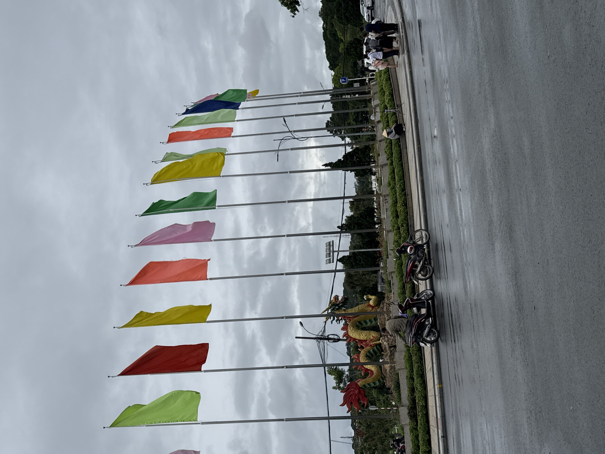 The image shows a street scene in Phường Xuân Hương - Đà Lạt, Vietnam. Along the side of the road, a row of tall metal flagpoles holds rectangular, solid-colored flags of green, red, yellow, orange, pink, blue, and other colors. To the right of the flags, along the edge of the road, is a low hedge separating the street from a small park area with a large, golden and red dragon sculpture. Parked motorbikes are visible next to the sculpture. Across the street, a group of people is standing near a parking sign. The sky is overcast and gray. The road appears to be wet, possibly from recent rain.