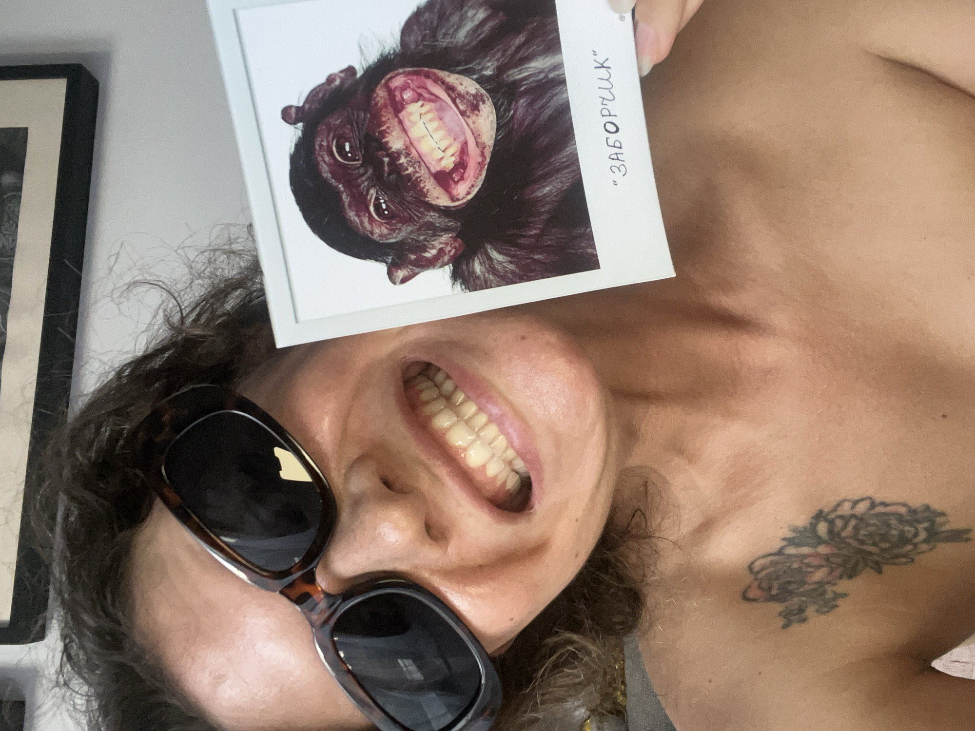 A person is holding up a photo of a chimpanzee with its mouth open wide, displaying its teeth. The person is wearing sunglasses and also has their mouth open, displaying their teeth. The photo is oriented so that both the person and the chimpanzee appear to be mimicking each other's expressions. There is a tattoo visible on the person's chest. The background is blurry, but a framed picture can be seen on the left side. There is no evidence of a location in the city of North Kuta, Indonesia.