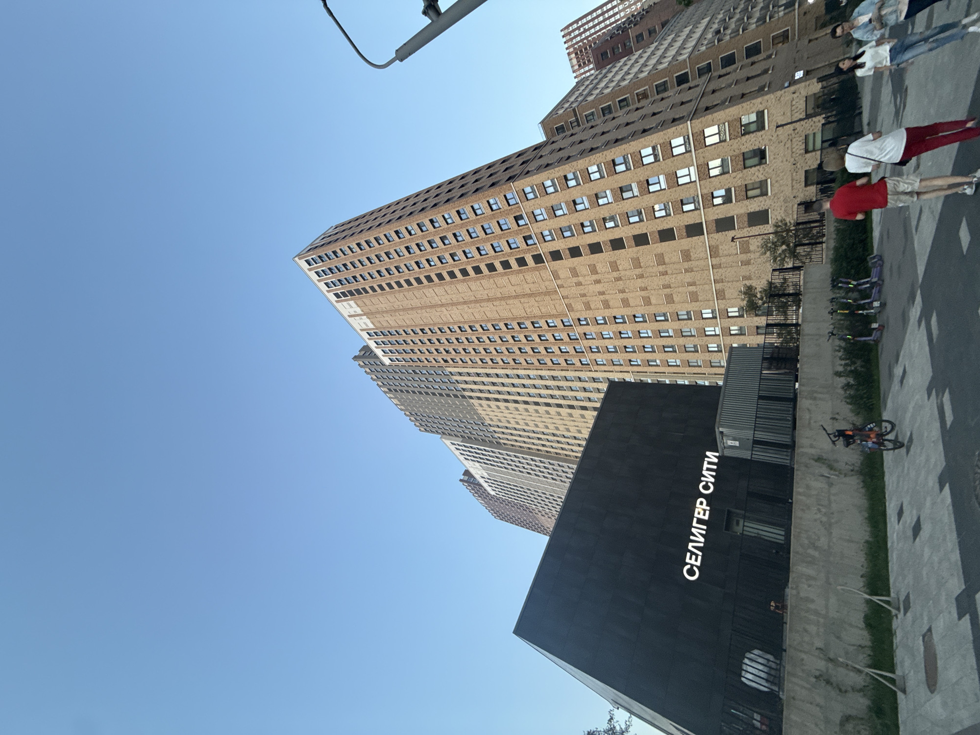 The image depicts a view of the "Seliger City" complex in Moscow, Russia. Tall buildings with repetitive window patterns dominate the background, reaching high into a clear blue sky. In the foreground is a dark, angular building with the name "СЕЛИГЕР СИТИ" (SELIGER CITY) written in white, Cyrillic letters on its side. Pedestrians can be seen walking on the pavement adjacent to the buildings. Bicycles and scooters are parked in the area. The camera angle points upwards, emphasizing the height of the buildings.