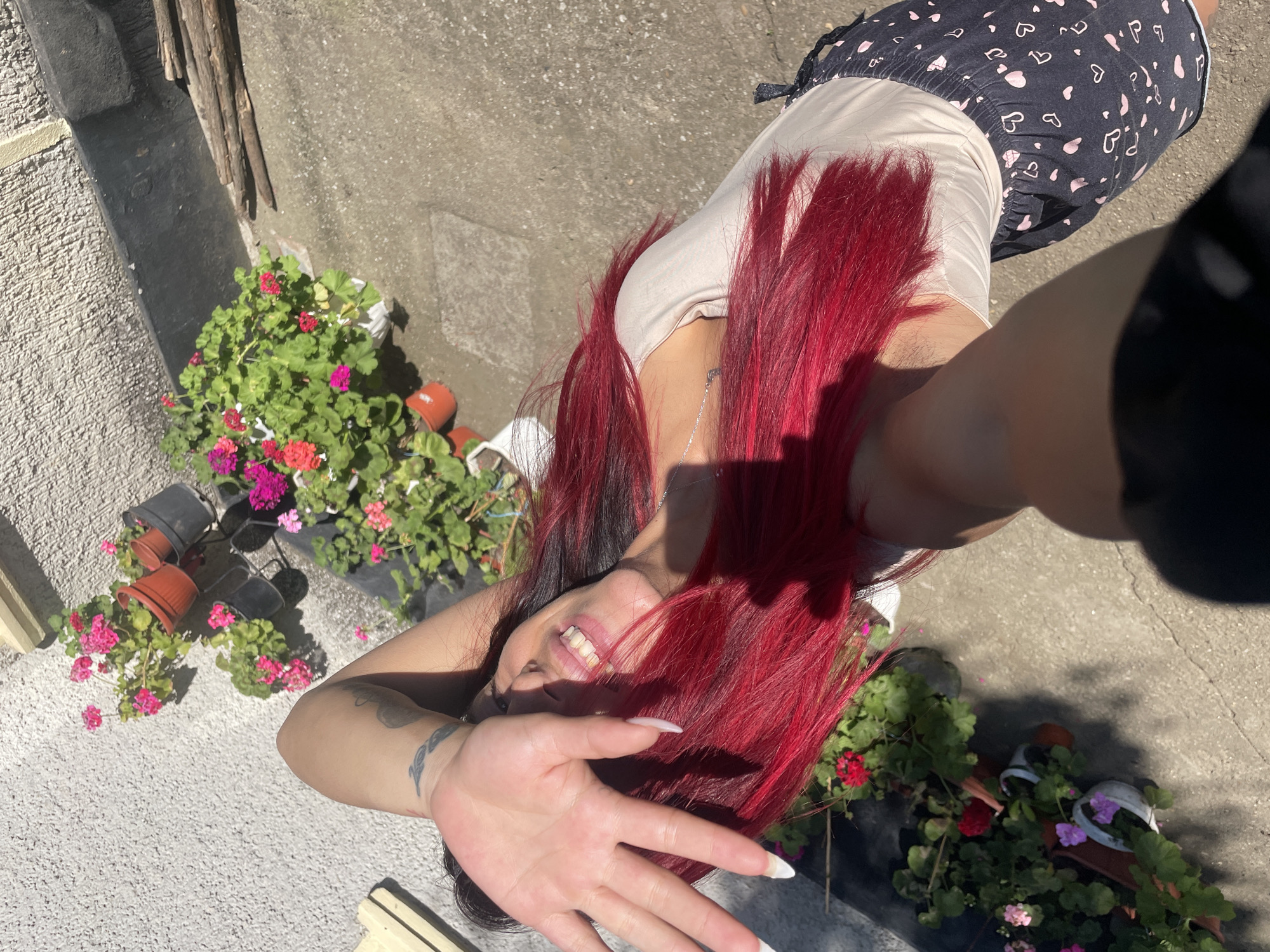An outdoor, high-angle, full-body shot captures a young woman with long, vibrant red hair posing in what appears to be a residential area, possibly Zheglartsi, Bulgaria. The woman is seen from the chest up, with her left arm extended and hand covering part of her face as she looks towards the camera with a wide smile. She is wearing a light-colored t-shirt and dark shorts patterned with small pink hearts. Her right arm is bent, with her hand resting on her head, partially obscuring her face. Behind her, a wall with a textured surface is visible, along with potted plants displaying bright pink and red flowers. The ground is paved with concrete, and a pile of wooden sticks lies near the wall. The scene is illuminated by bright sunlight, casting shadows.