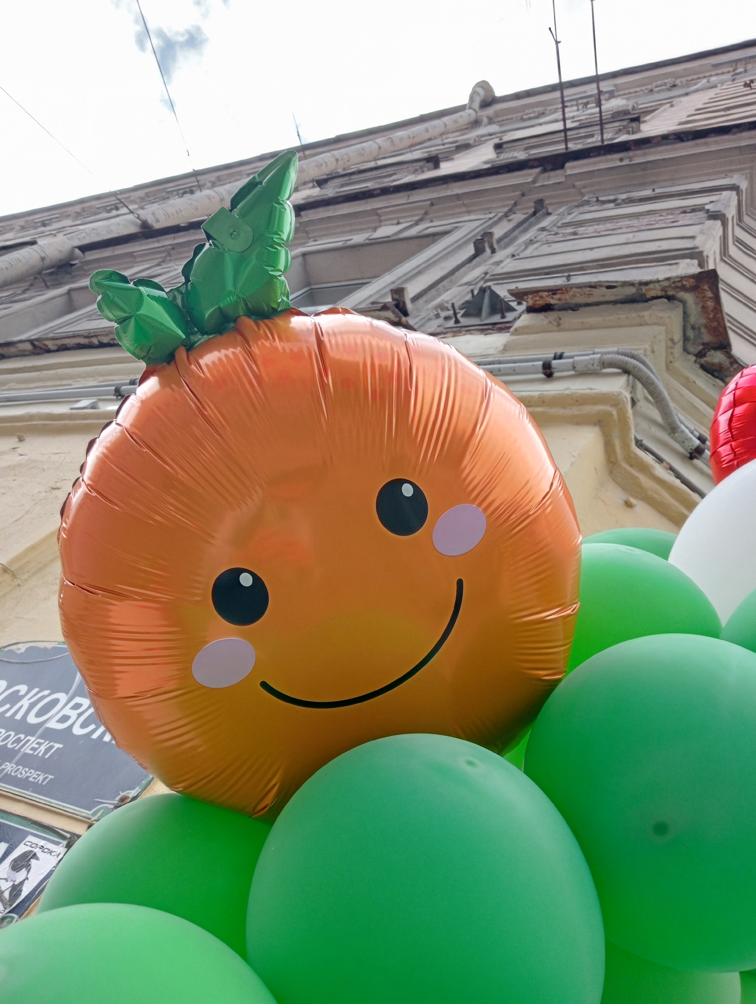 An orange carrot-shaped balloon with a smiling face and green leaves on top is the main subject of this image, situated outdoors against a backdrop of an aged, ornate building in Saint Petersburg, Russia. The balloon is surrounded by several green balloons and partially obscured by a white balloon and a red balloon, suggesting a festive or decorative arrangement. The building's architecture, with its detailed cornices and weathered facade, indicates an older part of the city. The sky above is overcast with visible clouds. Signs with Cyrillic text, including "МОСКОВСКИЙ ПРОСПЕКТ" (Moskovsky Prospekt), are visible in the lower left corner, confirming the location as Moskovsky Prospekt in Saint Petersburg. The image is taken from a low angle, looking upwards towards the balloon and the building.