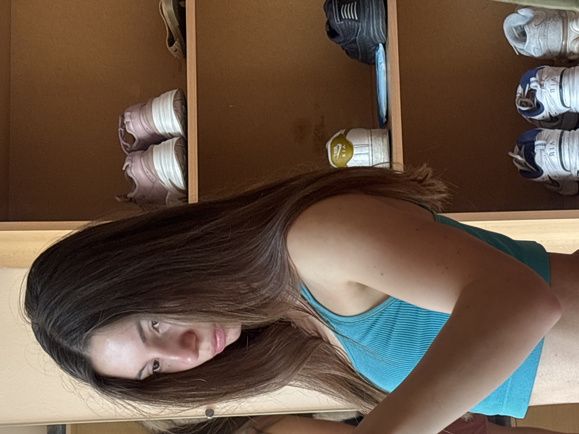 The image shows a young woman in a teal crop top, with long brown hair, looking down. She is standing in what appears to be a closet or a room with shelves filled with shoes. The shelves are made of wood, and there are several pairs of sneakers and other footwear on them. In the background, a mirror reflects part of the room. The lighting suggests it is daytime. The location is described as being in Varna, Bulgaria.