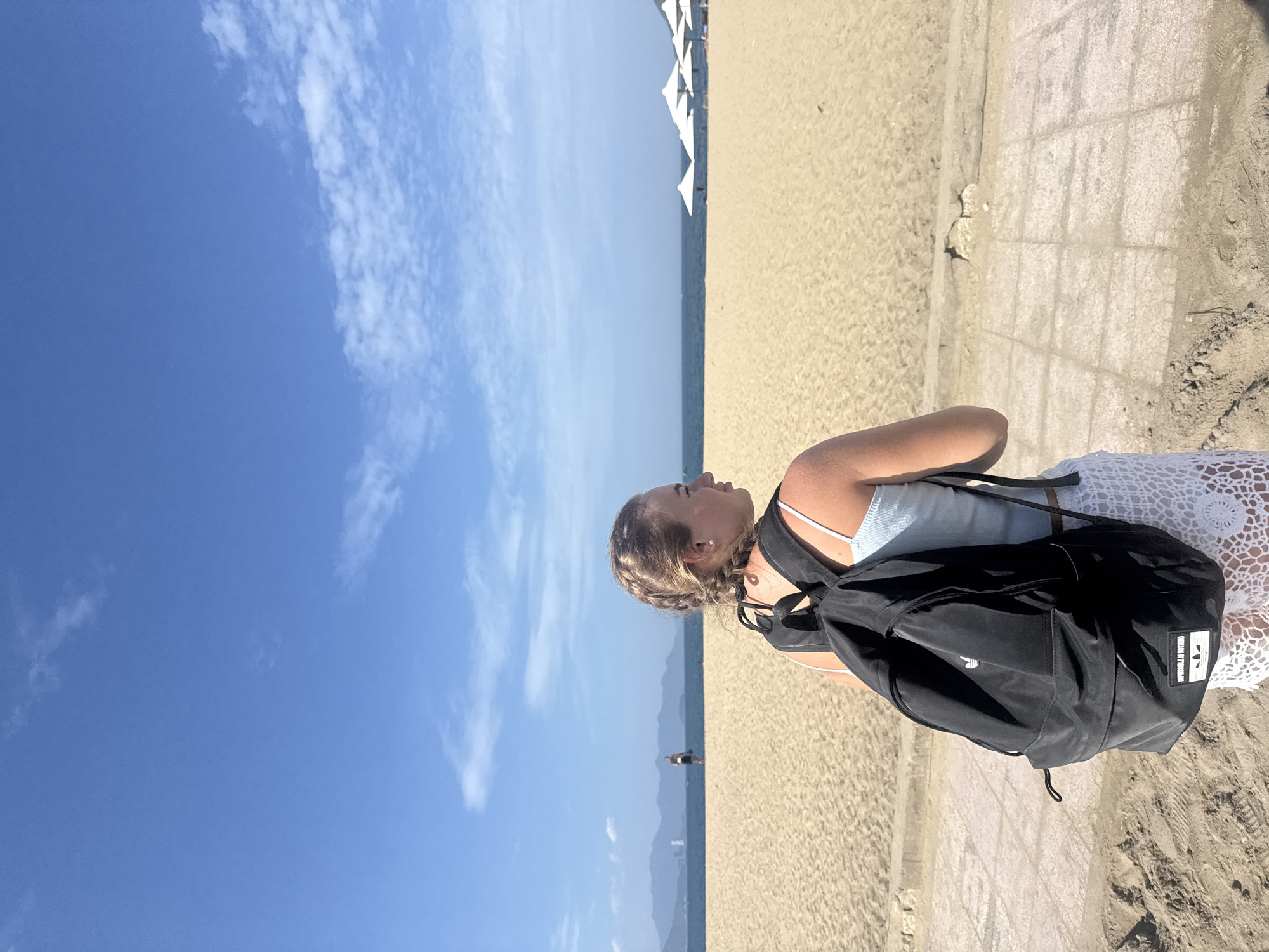 A young Caucasian woman with blonde hair in braids, wearing a light blue tank top and a white crochet skirt, stands facing away from the camera on a paved pathway next to a sandy beach. She has a black backpack on her shoulders. The beach is wide and empty, with the ocean in the background. Several white umbrellas are set up on the sand. In the distance, mountains can be seen. The sky is blue with scattered white clouds. The location is likely Tran Phu Beach in Nha Trang, Vietnam, a popular tourist destination known for its long sandy beaches and coastal promenade.