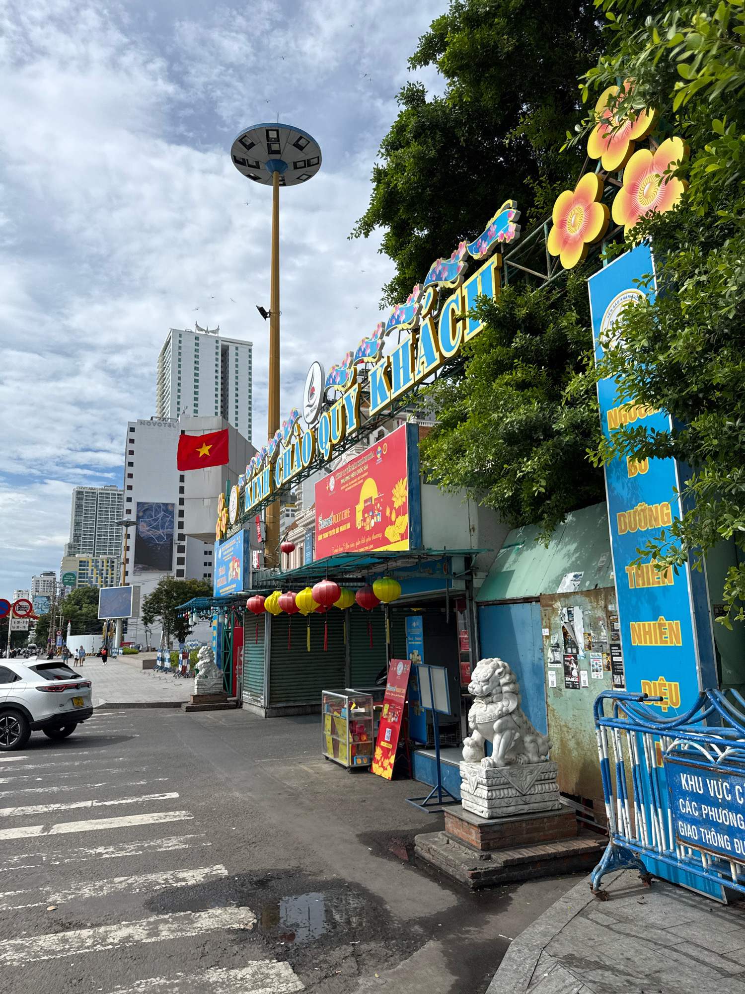This is a street-level, eye-level, medium shot of a closed shopfront and street scene in Nha Trang, Vietnam. The shopfront is decorated with a large, blue and yellow sign with Vietnamese text that reads "KINH CHAO QUY KHACH". Below the sign, red and yellow lanterns are hung. To the left of the shopfront, a white car is parked on the street, and a crosswalk is visible. A tall, modern building with a Vietnamese flag is in the background. To the right of the shopfront, a blue barrier fence with white stripes and Vietnamese text is present. A stone lion statue stands in front of the shop. The sky is cloudy with a few birds flying.