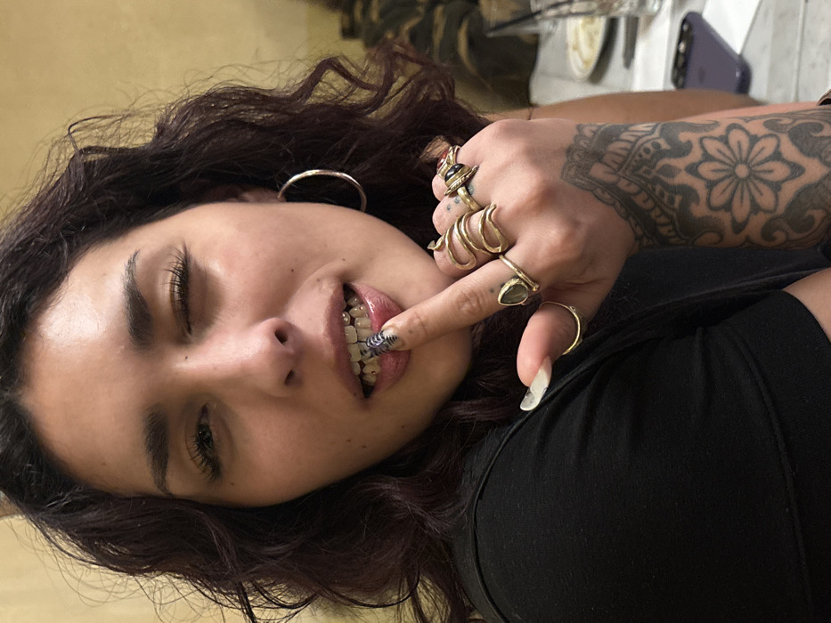 A young adult female is depicted in a medium close-up, looking directly at the viewer from an indoor setting within the city of Paris, France. She has dark, wavy hair and wears a black short-sleeved top and a large silver-toned hoop earring in her right ear. Her left eye is closed in a wink, and her lips are slightly parted, revealing upper teeth adorned with small, reflective tooth gems or grillz. The index finger of her right hand is brought to her lower lip; this finger features a long, pointed fingernail painted with a black and white striped pattern. Her right hand is heavily adorned with multiple gold-toned rings: a prominent snake-shaped ring on her middle finger, a large ring with a dark stone on her index finger, and several other rings on her ring and pinky fingers. A detailed black and grey tattoo, featuring a mandala-style design with a central flower motif, covers her right forearm. The background is blurred, showing elements consistent with a commercial establishment such as a restaurant or cafe, including a faint yellow wall, a table with white plates, a glass containing a dark beverage and a straw, and a purple mobile phone. The specific location within Paris is not visually identifiable.