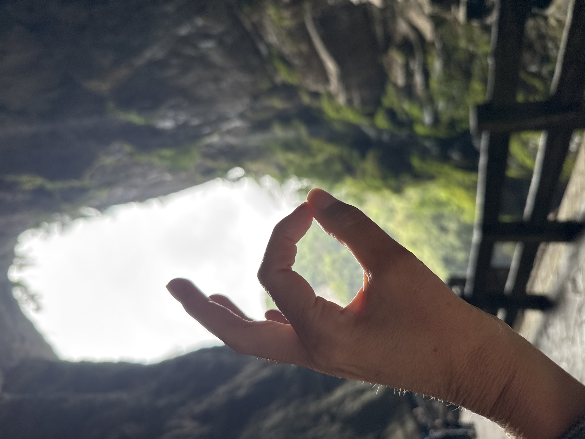 The image shows a hand in the Gyan mudra gesture with a blurred background of a cave-like opening with natural light. The location is Wulong District, China, as the image is taken from the Three Natural Bridges area, known for its caves and karst landscapes. A wooden walkway is partially visible to the right.