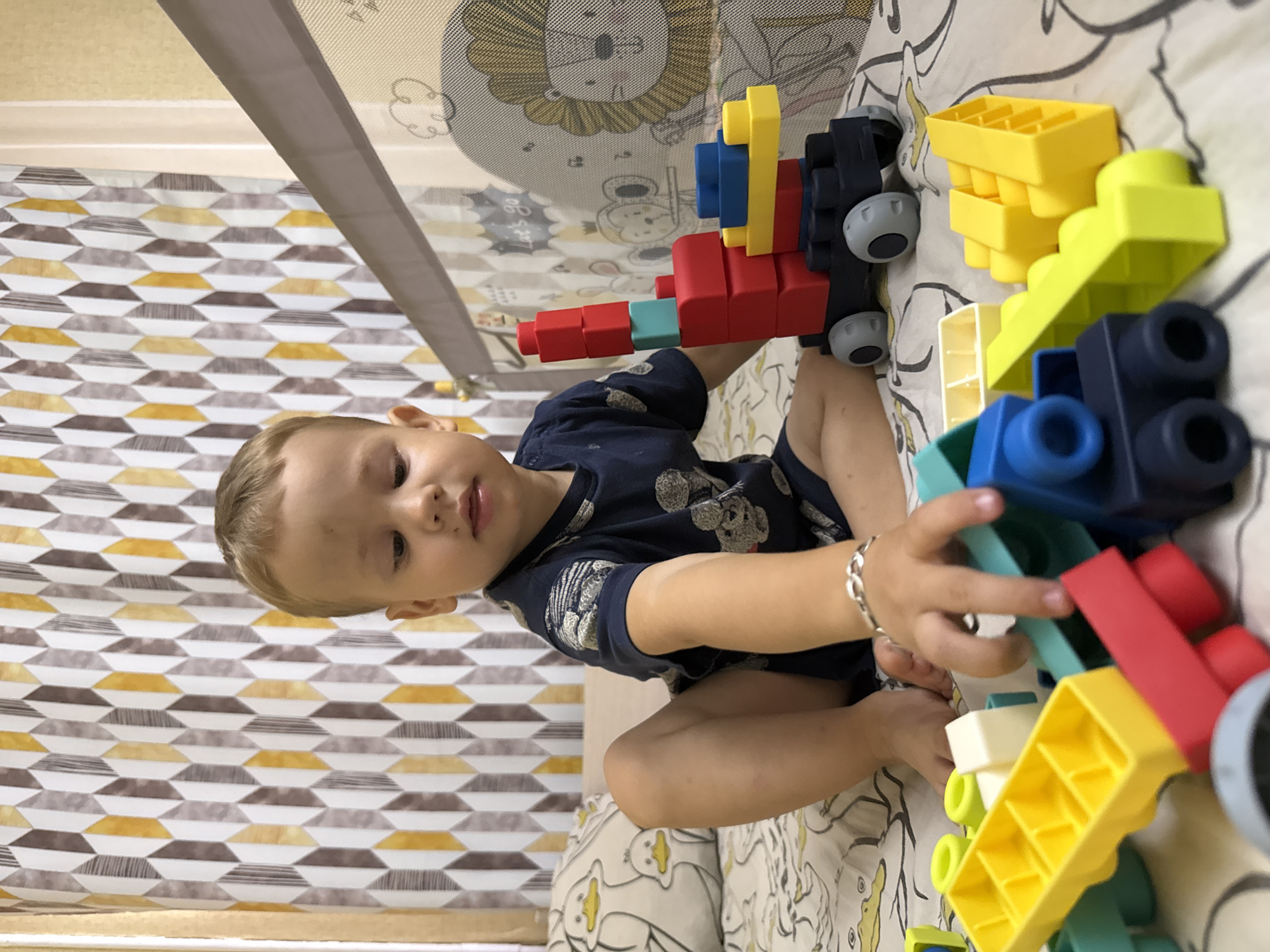 A young child, approximately two years old, sits on a bed in what appears to be a nursery or playroom. The child, with fair skin and light brown hair, is wearing a dark blue t-shirt with a pattern of cartoon characters. The child's legs are spread apart, and their arms are reaching towards a collection of colorful building blocks. The blocks are made of plastic and come in various shapes and colors, including red, blue, yellow, and green. One block structure resembles a crane, with a red base, a blue platform, and a stack of red and teal blocks forming the boom. The bed is covered with a white and yellow patterned sheet, featuring duck illustrations. In the background, a grey crib with a mesh side is visible, adorned with cartoon graphics of animals and text. A curtain with a geometric pattern of yellow, brown, and white rhombuses hangs behind the child. The overall setting suggests a domestic environment in Rusanovo, Russia, likely a child's bedroom or play area.