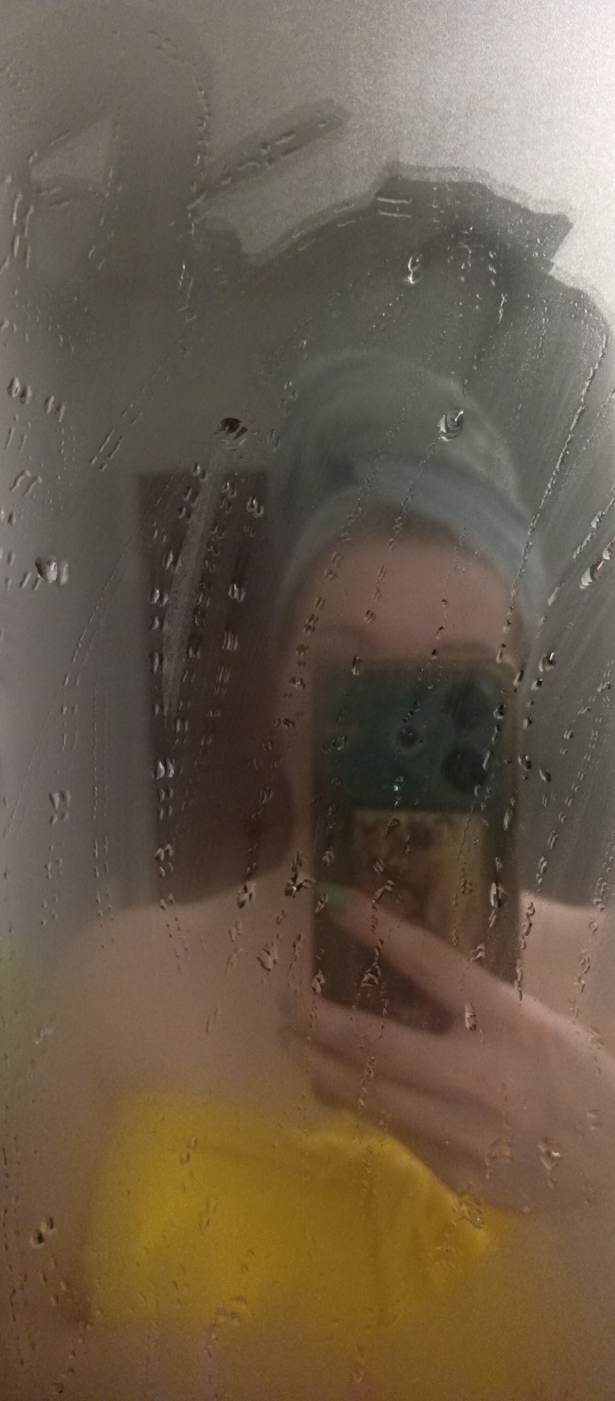 A person is reflected in a fogged or wet mirror. The person has a towel wrapped around their head and is holding a smartphone in their hand. A yellow garment is visible. The surrounding environment is blurry due to the condensation. The city location is Unknown, Unknown.