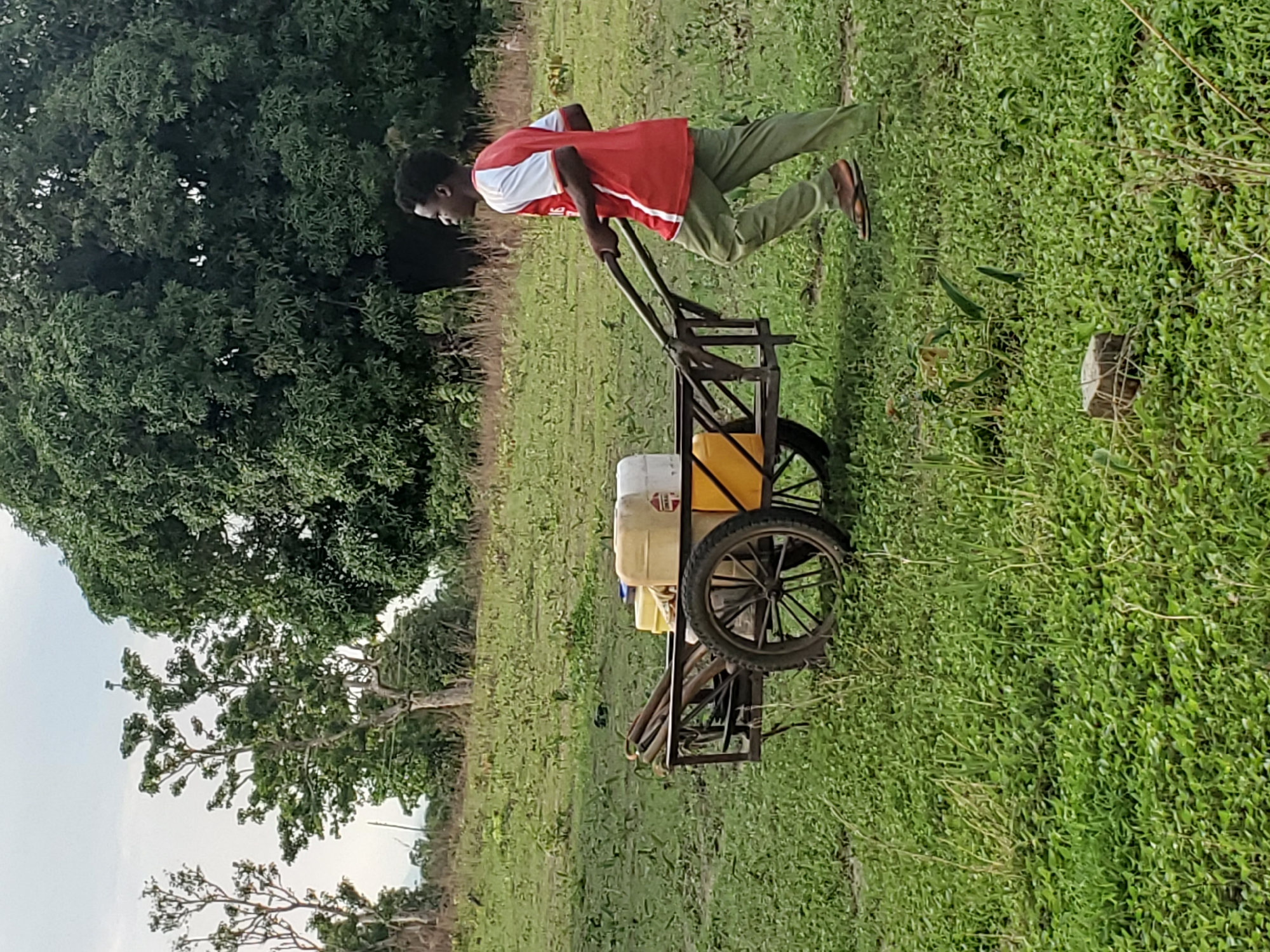 In an outdoor scene in Mararaba, Nigeria, a young man pulls a wheeled cart across a grassy field. He wears a red and white short-sleeved shirt, green pants, and sandals. The cart is a metal frame with two large wheels and carries several yellow and white containers. The environment consists of green grass, trees, and a cloudy sky.