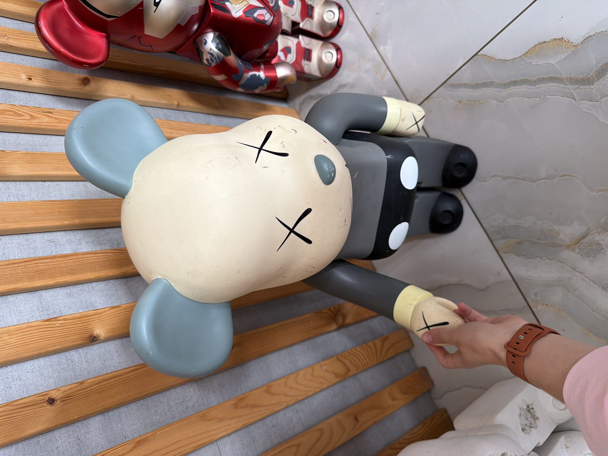 A person's arm and hand is visible in the lower left of the frame, wearing a brown watch and a pink shirt sleeve. The hand is reaching out to touch the foot of a large, gray and cream-colored toy figure with blue ears and black X's for eyes. Behind the toy figure is a red and silver toy figure. The figures are standing on a marble-patterned floor, in front of a wall made of vertical wooden planks and gray paneling. The scene is indoors, likely in a residential setting or shop in the city of Semiluki, Russia.