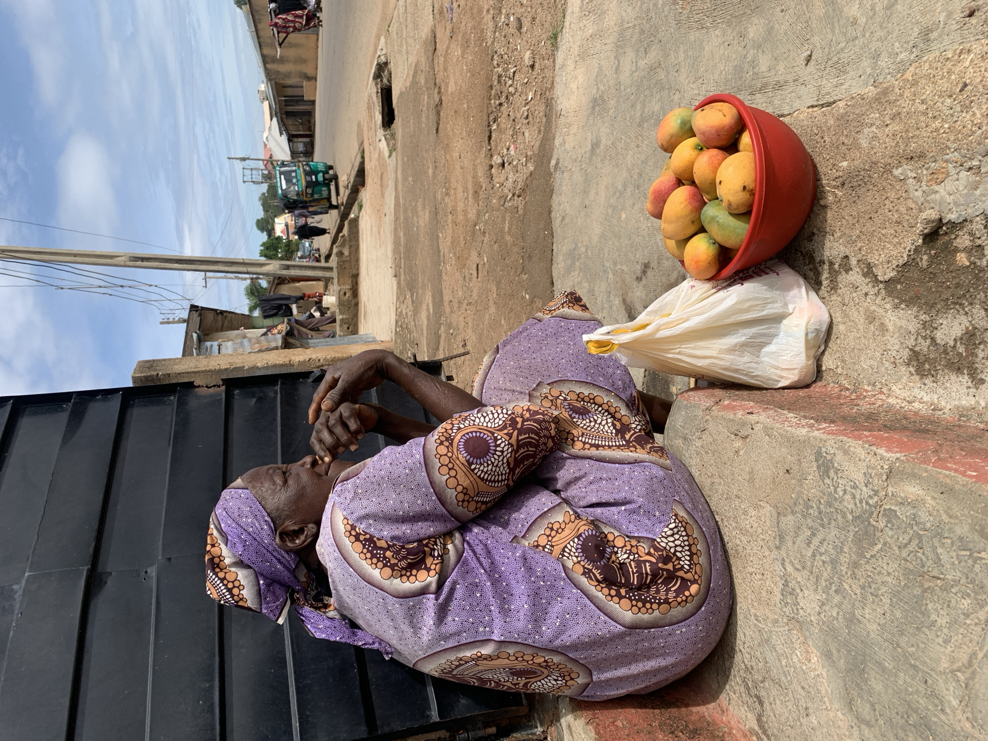 An elderly dark-skinned woman is seated on concrete steps in Jos, Nigeria, looking right. She wears a purple headwrap and a matching dress adorned with brown and white circular patterns. Her hands are clasped together near her mouth. To her immediate right, on the ground, a white plastic bag with yellow handles rests beside a red plastic bowl containing approximately 10-15 mangoes of varying ripeness (yellow, orange, green, red). Behind the woman is a black corrugated metal wall. The background shows a dirt and concrete street with utility poles and overhead wires. Buildings line both sides of the street. A green and yellow motorized tricycle, marked "GIFT FROM GOD" and "104," is visible on the street, moving away from the viewer. Another figure walks near the tricycle. The sky above is partly cloudy, indicating daytime.