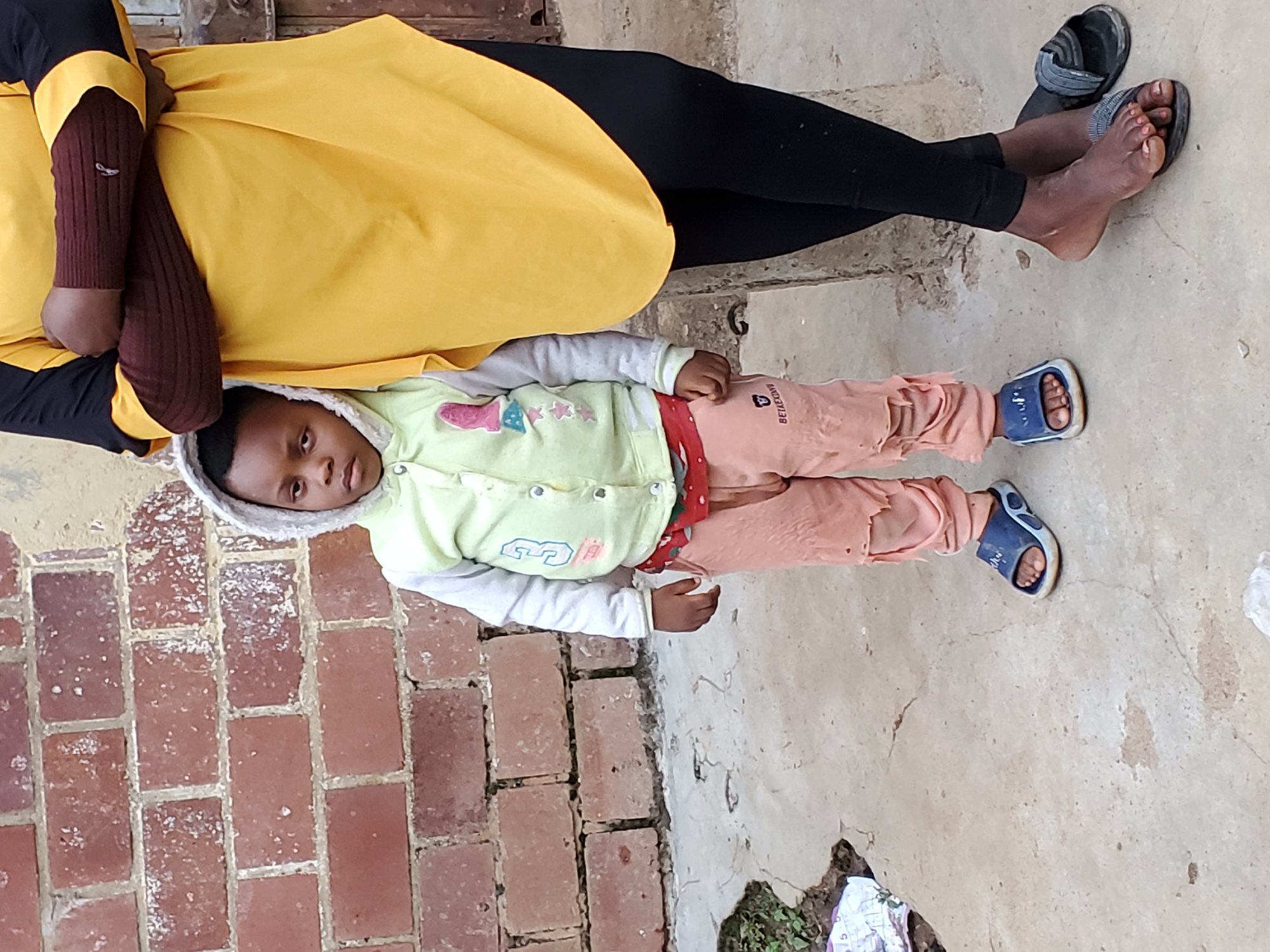 A child stands on a concrete surface in front of a brick wall. The child is wearing a light green hooded jacket over a grey sweater, light pink pants with ripped knees, and blue slide sandals. To the right of the child, an adult's legs are visible, wearing black leggings and black slide sandals. The adult's arms are crossed. The scene appears to be outdoors, likely in an urban setting like Jos, Nigeria, given the visible brickwork and concrete ground.