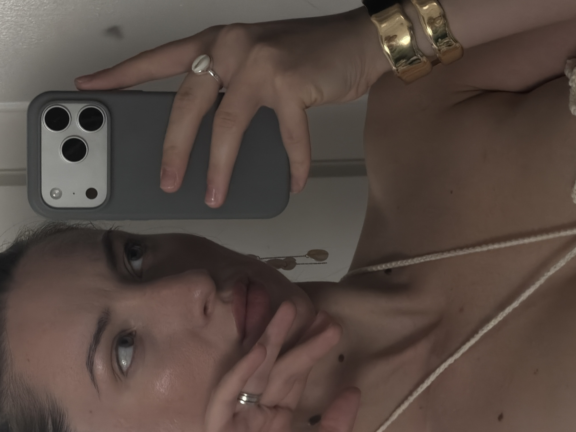 A woman is taking a selfie in a well-lit room, possibly a hotel room in Sharm El Sheikh, Egypt. She is holding a gray phone with multiple cameras to her right and looking at the phone's screen. The woman has her left hand near her face, with a ring on her ring finger and another ring on her middle finger. She is wearing large, gold cuff bracelets on her right wrist and a necklace with a white cord. The background is plain and appears to be a wall.