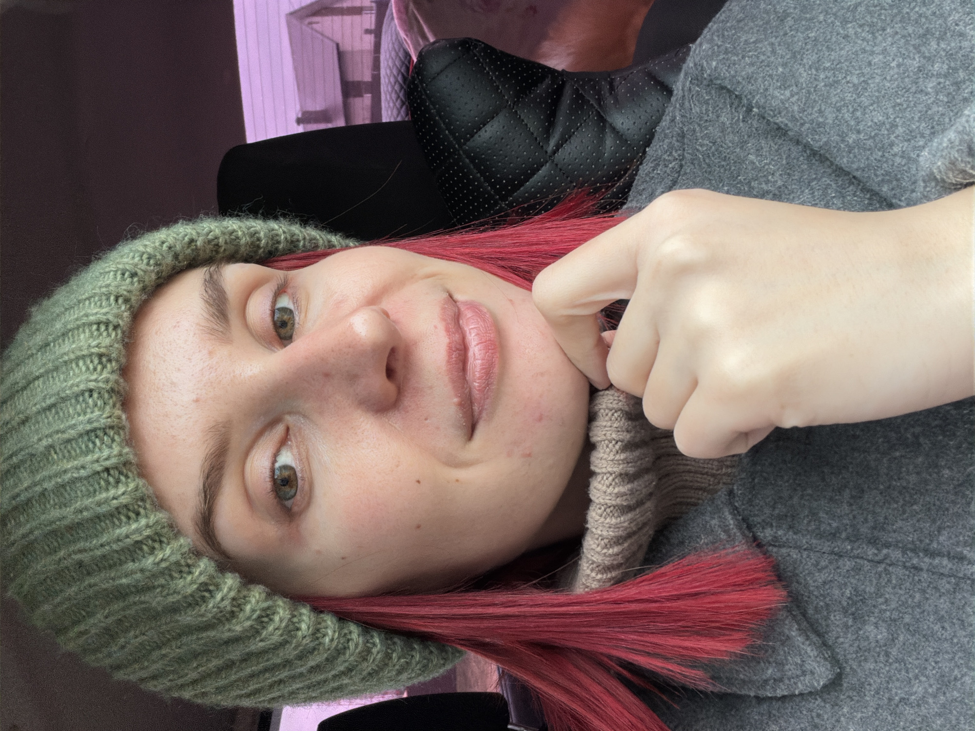A close-up selfie shows a person with fair skin, green eyes, and reddish-pink hair, positioned diagonally across the frame. They are wearing a green knitted beanie, a beige knitted turtleneck or sweater, and a grey woolen coat or jacket, suggesting cool weather. The person looks directly at the camera with a subtle, pleasant expression, while their left hand gently pulls at their lower cheek/chin. Visible on their skin are a few minor blemishes.
The background indicates an indoor setting, likely within a vehicle, as a black quilted car seat or headrest is visible behind them. Through a window in the upper left, a blurry light purple or pink structure can be seen, possibly part of a building. The image is taken in what appears to be daytime conditions. No text is visible in the image. The location context provided is Chaikovsky, Russia.