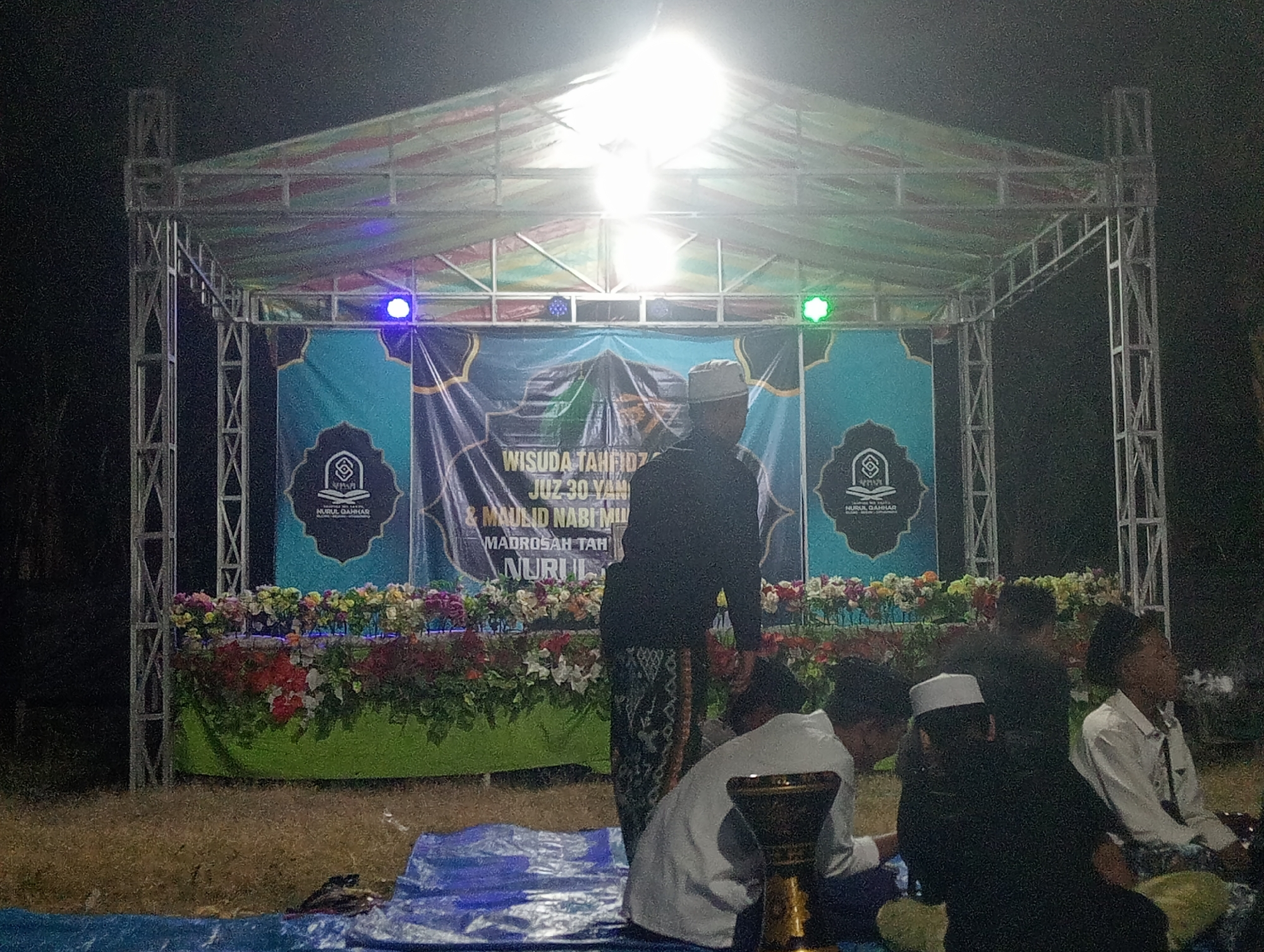 The image captures an outdoor evening event at night, possibly a graduation or religious ceremony, in Bloro, Indonesia. A temporary stage is erected, covered by a multi-colored tarp and illuminated by bright overhead lights and colored spotlights (blue and green). A large banner with Arabic script and Islamic motifs hangs as a backdrop, featuring text that appears to read "WISUDA TAHFI" and "MAULID NABI MUH".
In front of the banner, a flower arrangement lines the stage. Several people are visible, some standing and others seated on blue tarps on the ground in front of the stage. One person in the foreground, facing away from the camera, is wearing traditional Indonesian attire, including a batik sarong and a peci hat. Another person in the middle ground, also facing away, is dressed in a black shirt and sarong, and is wearing a white peci. Others are partially obscured or in the background. The ground appears to be a mix of dry grass and earth, with blue tarps laid out for seating. The overall atmosphere suggests a community gathering under festive illumination.