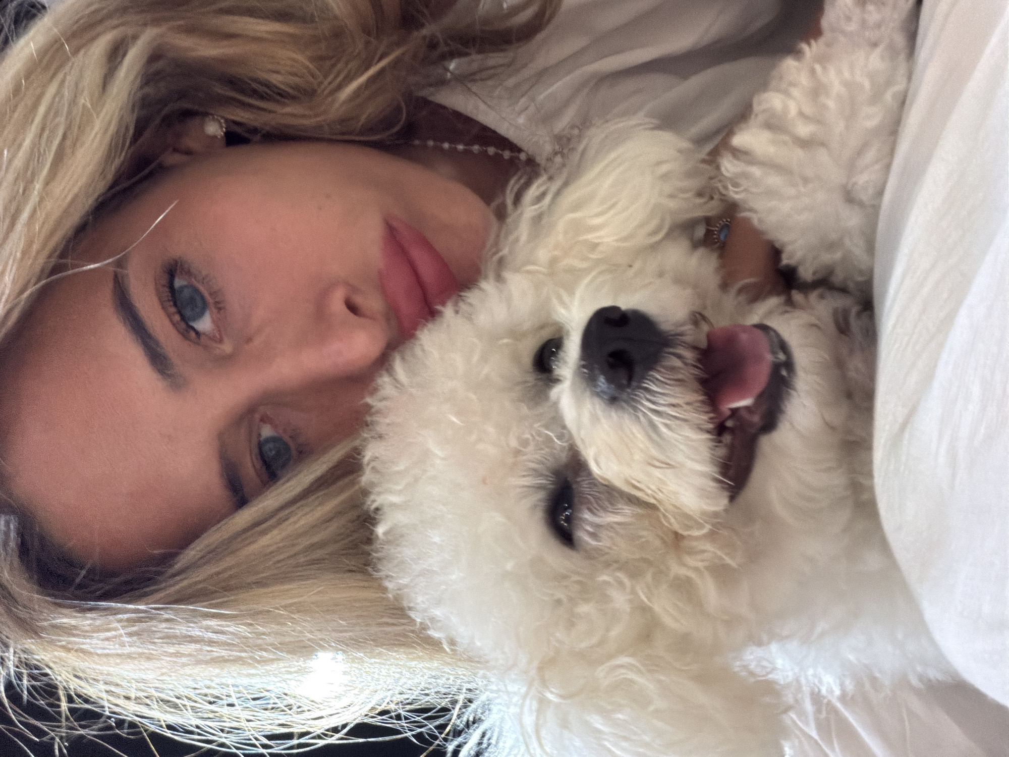 The image shows a woman and a small, white dog lying close together. The woman has blonde hair, fair skin, and blue eyes. Her head is tilted, and she is looking towards the camera. The dog, possibly a Bichon Frise or similar breed, is positioned next to her face with its tongue hanging out. The scene appears to be indoors, potentially in bed, with a white sheet visible. The location is not determinable as being in Kedewatan, Indonesia.