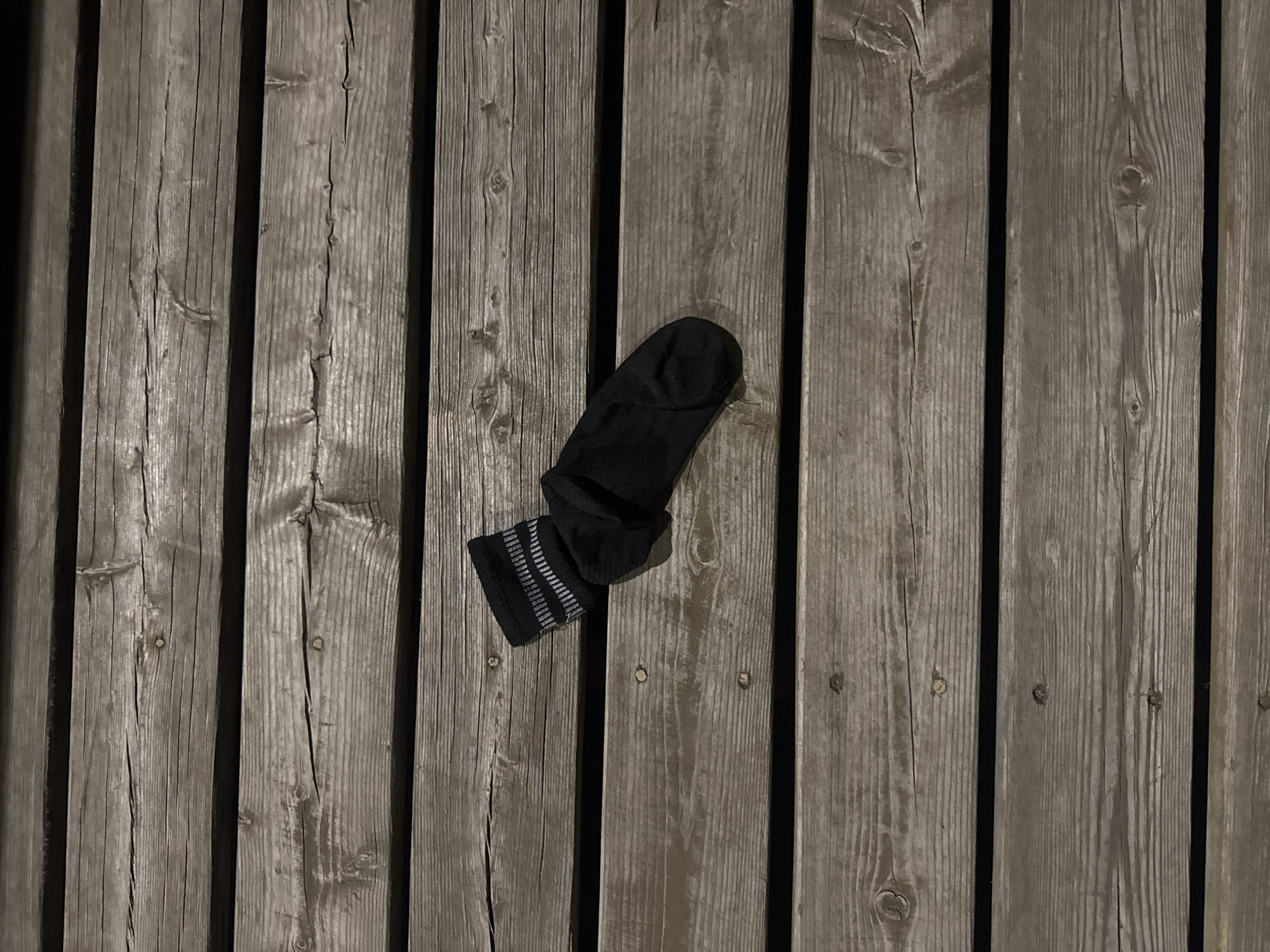 A single black sock lies on a gray wooden plank surface. The sock has gray stripes around the cuff. The planks are vertically oriented, with noticeable gaps between them. The image appears to be taken at night or in low lighting conditions. There are no people visible in the image. The location is not identifiable from the image.
