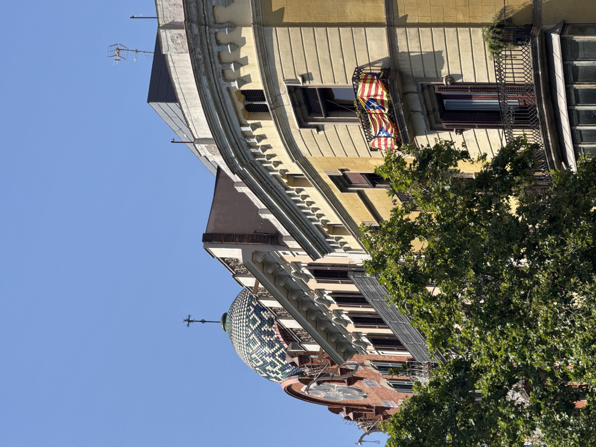 The image captures a sunlit view of the facade of a historic building in Barcelona, Spain. The building exhibits distinctive architecture, featuring a green and white tiled dome topped with a cross, and ornate balconies with intricate ironwork. A Catalan flag is prominently displayed on one of the balconies. Lush green trees partially obscure the lower portion of the building. The clear blue sky suggests a bright, daytime setting.