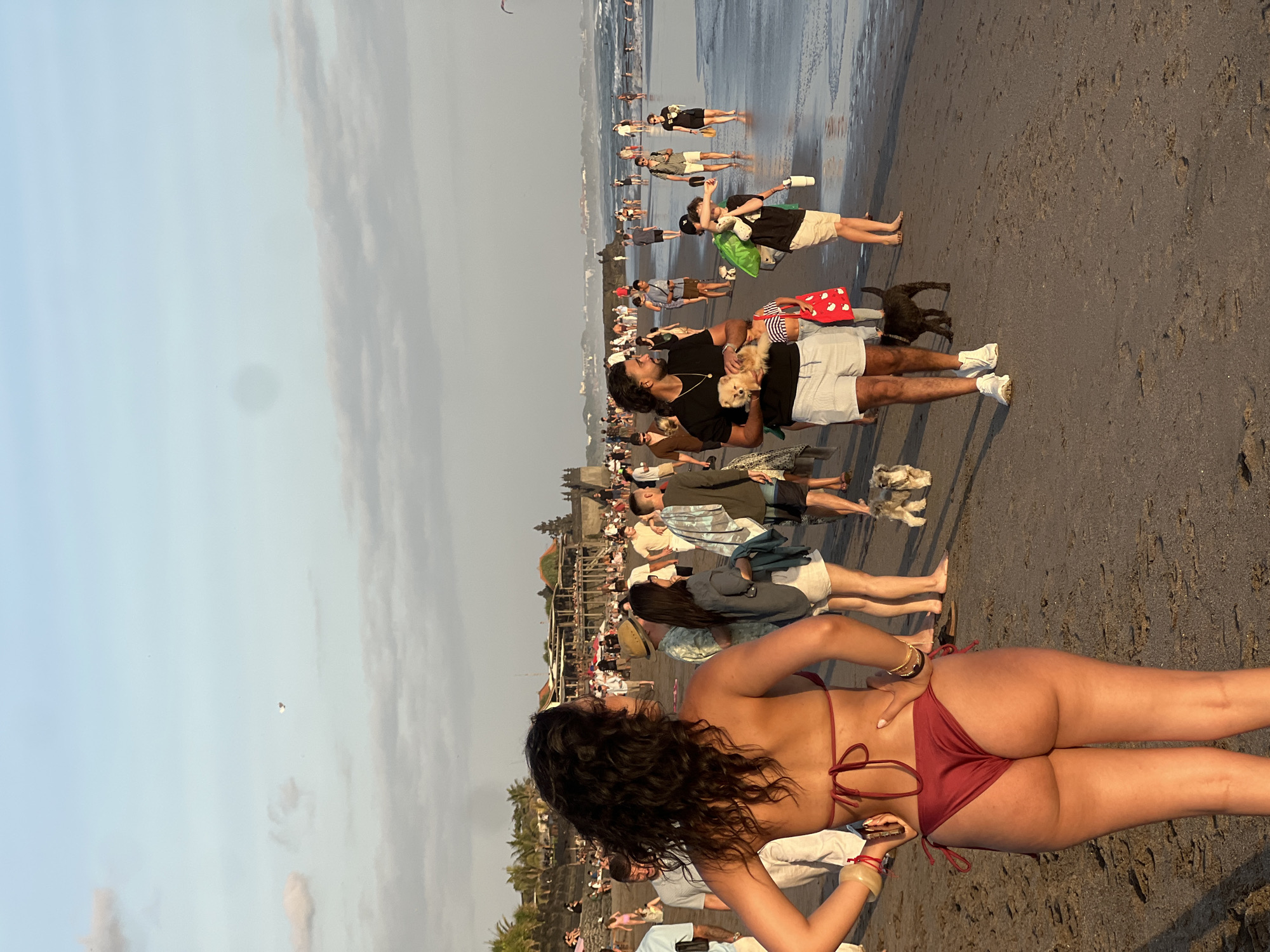 An overhead drone shot captures a wide view of a crowded beach in North Kuta, Indonesia, bathed in the warm glow of late afternoon sun. The sandy shore stretches out, dotted with numerous people enjoying the beach. In the foreground, a woman wearing a red bikini faces away from the camera, her hands on her hips. A variety of individuals are visible in the midground, some walking, others gathered in small groups, and a few walking dogs. The ocean forms the backdrop, with gentle waves lapping at the shore and more people wading in the water further out. In the distance, a series of buildings with traditional Balinese architecture line the coastline, suggesting a developed resort area. The sky above is a soft blue with scattered clouds.