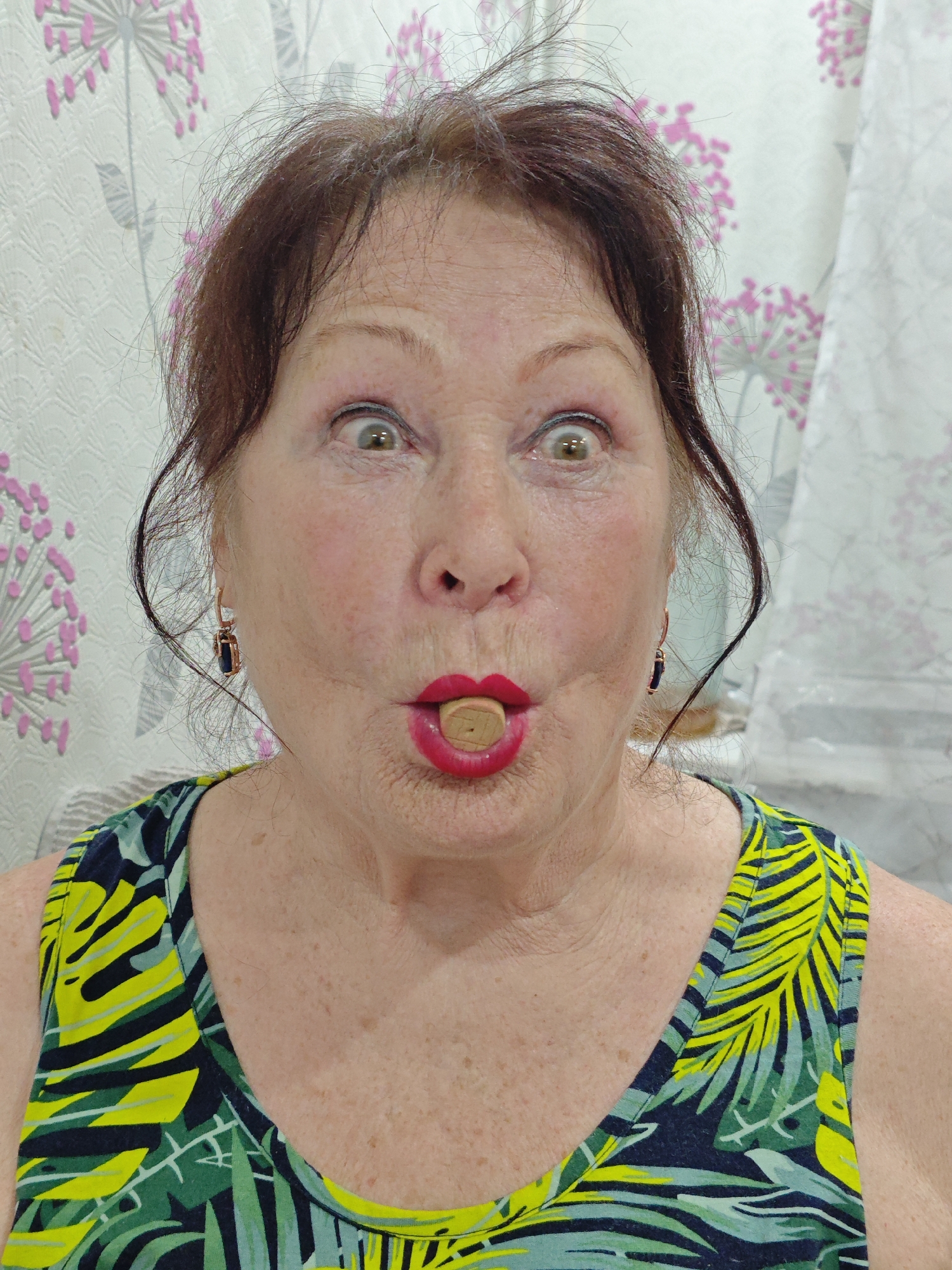 This indoor image, captured in Markovskii, Russia, features a single adult woman, appearing to be middle-aged to elderly, engaging in a playful pose. She has dark brown hair, styled back with some loose strands, and is wearing a sleeveless top with a vibrant tropical leaf pattern in shades of green, yellow, and dark blue. Her eyes are wide open, expressing an exaggerated sense of surprise or humor, while her mouth is puckered around a light-colored cylindrical object, which appears to be a cork or a similar wooden item. She wears red lipstick, which is clearly visible around the object. Gold-toned hoop earrings, each with a dark, rectangular stone, are visible on her earlobes. The background features a light-colored wallpaper adorned with a repeating pattern of stylized pink and gray dandelion-like flowers or seed heads. The lighting is bright and even, consistent with an indoor setting during daytime or under good artificial illumination. The woman's expressive face and the object in her mouth suggest a humorous or theatrical moment. No text is visible in the image.