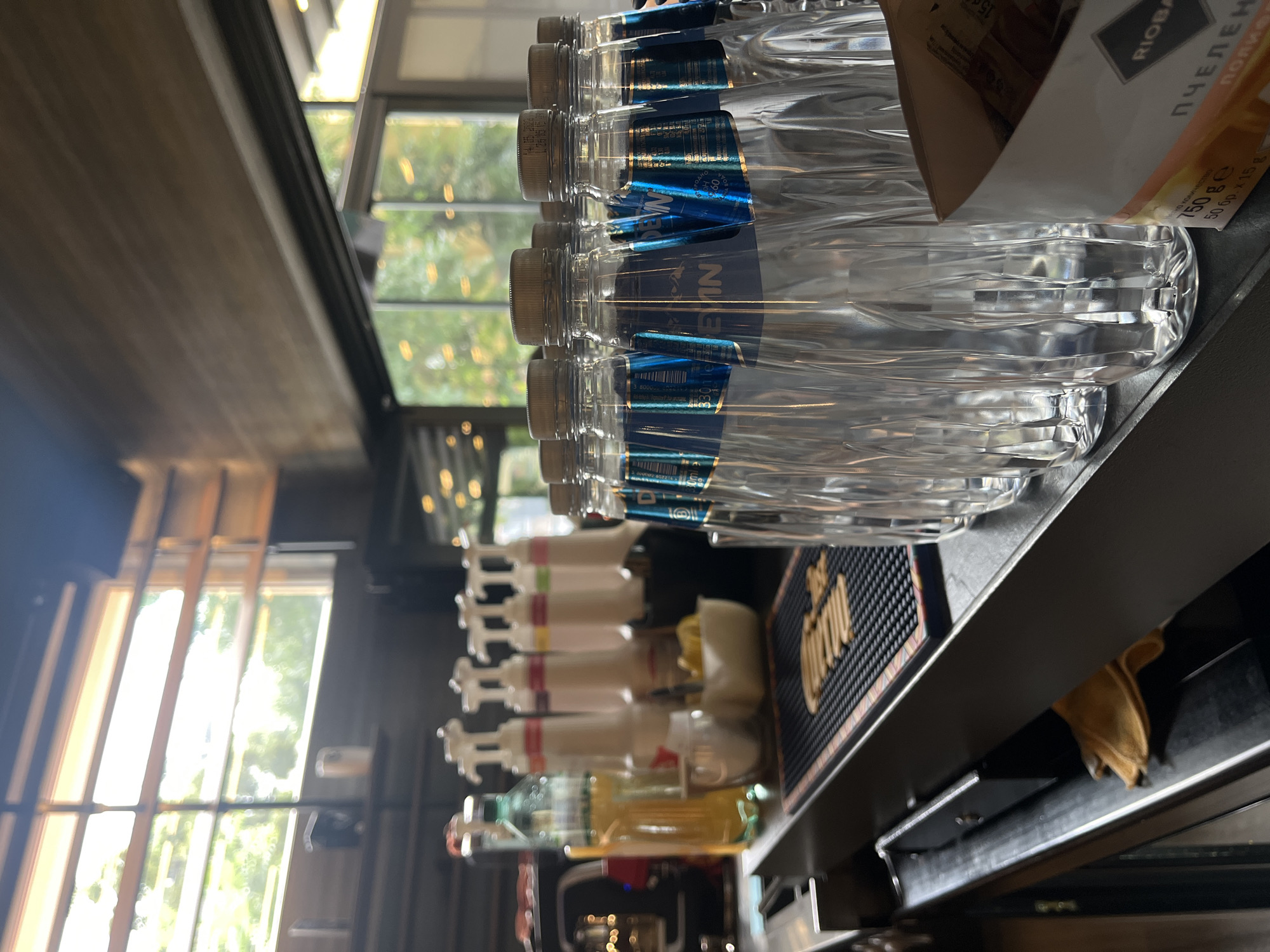 This is a photograph of a bar counter with several bottles of water and dispensers. The scene appears to be indoors, possibly a cafe or restaurant, with large windows in the background showing greenery. The bottles of water are clear plastic with blue labels, and there are multiple white dispensers with colored tops in the middle ground. A yellow bottle of liquid, possibly syrup or juice, is on the left. A black mat with a pattern is in front of the dispensers. In the foreground, on the right, is a box with orange and white packaging. The lighting suggests it is daytime. The image does not provide any explicit clues about its location in Plovdiv, Bulgaria. There are no people visible in the photograph.