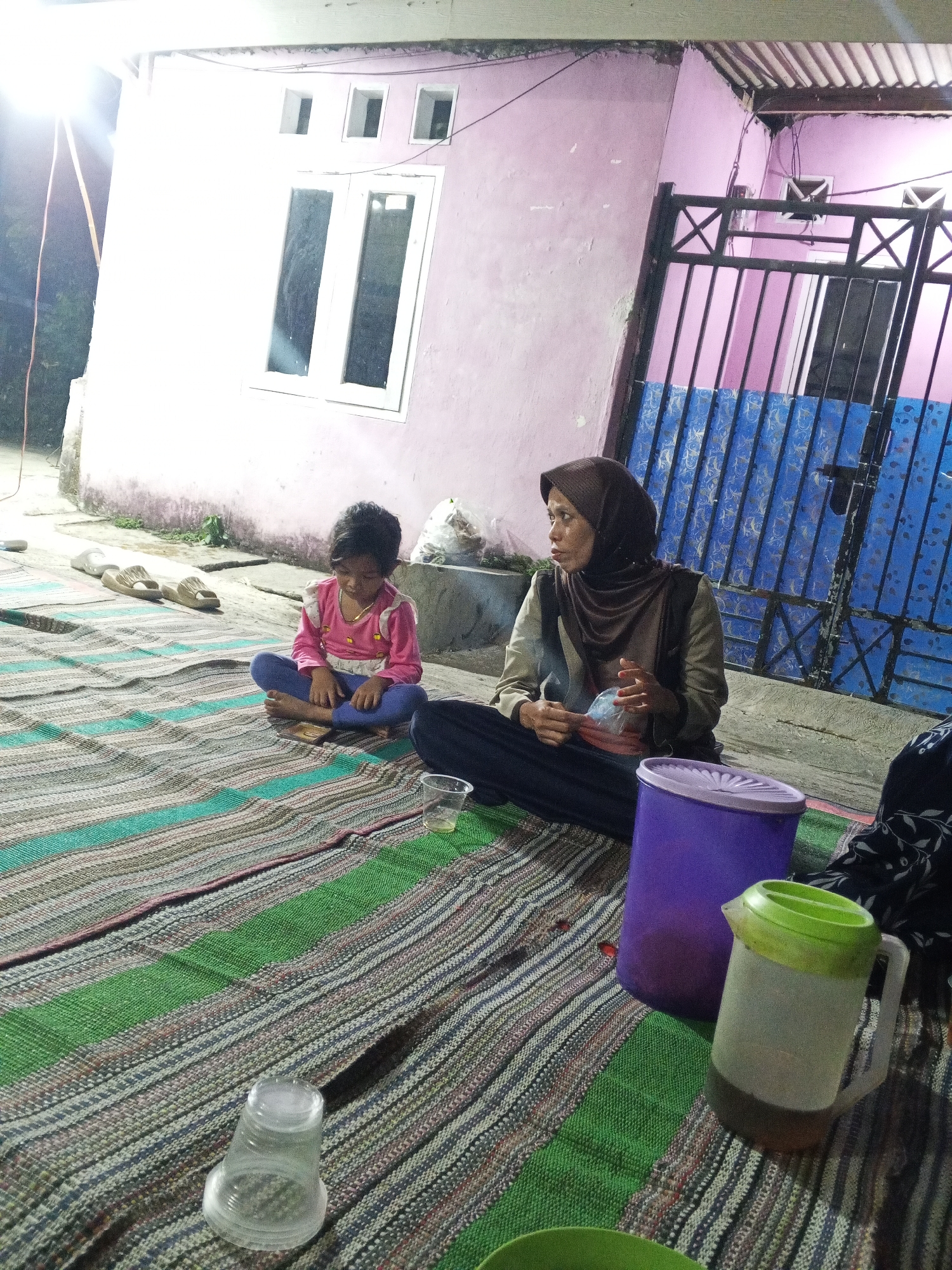 In Ciruas, Indonesia, a woman wearing a hijab and a neutral-toned shirt sits on a woven mat, gesturing with her hands while speaking. Beside her, a young child in a pink shirt and blue pants is seated cross-legged, looking down. A purple water dispenser and a pitcher with a green lid are placed on the mat near the woman. The scene appears to be outdoors, with a pink building and a black metal gate in the background. A bright light is visible to the left of the frame.