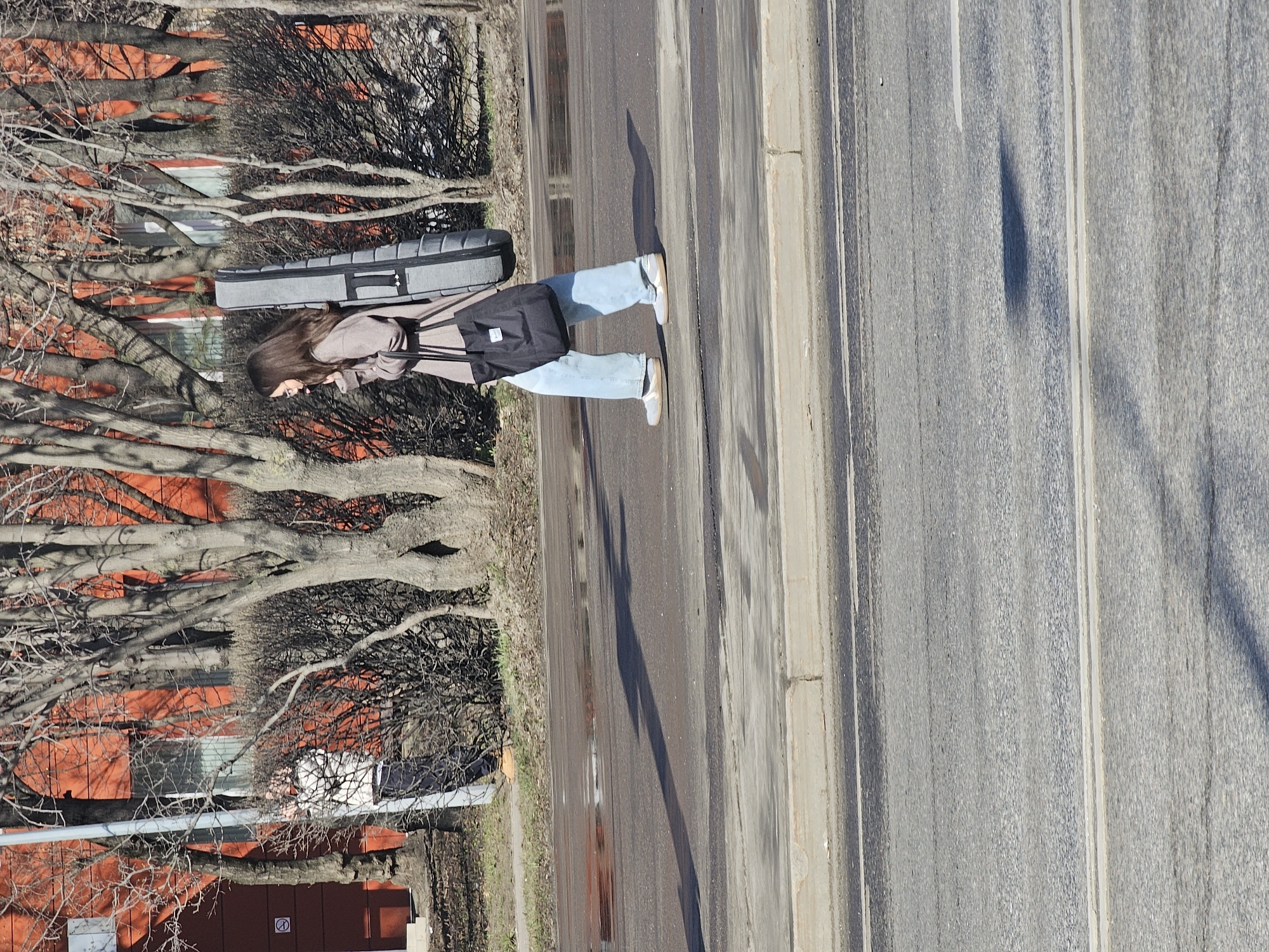 A person is standing on the edge of a road. They are wearing a grey jacket, light blue jeans, and carrying a black bag and a grey rectangular case. The road is grey with dividing lines and a concrete sidewalk to the right. To the person's left are trees and a red building. The location is Khimki, Russia.