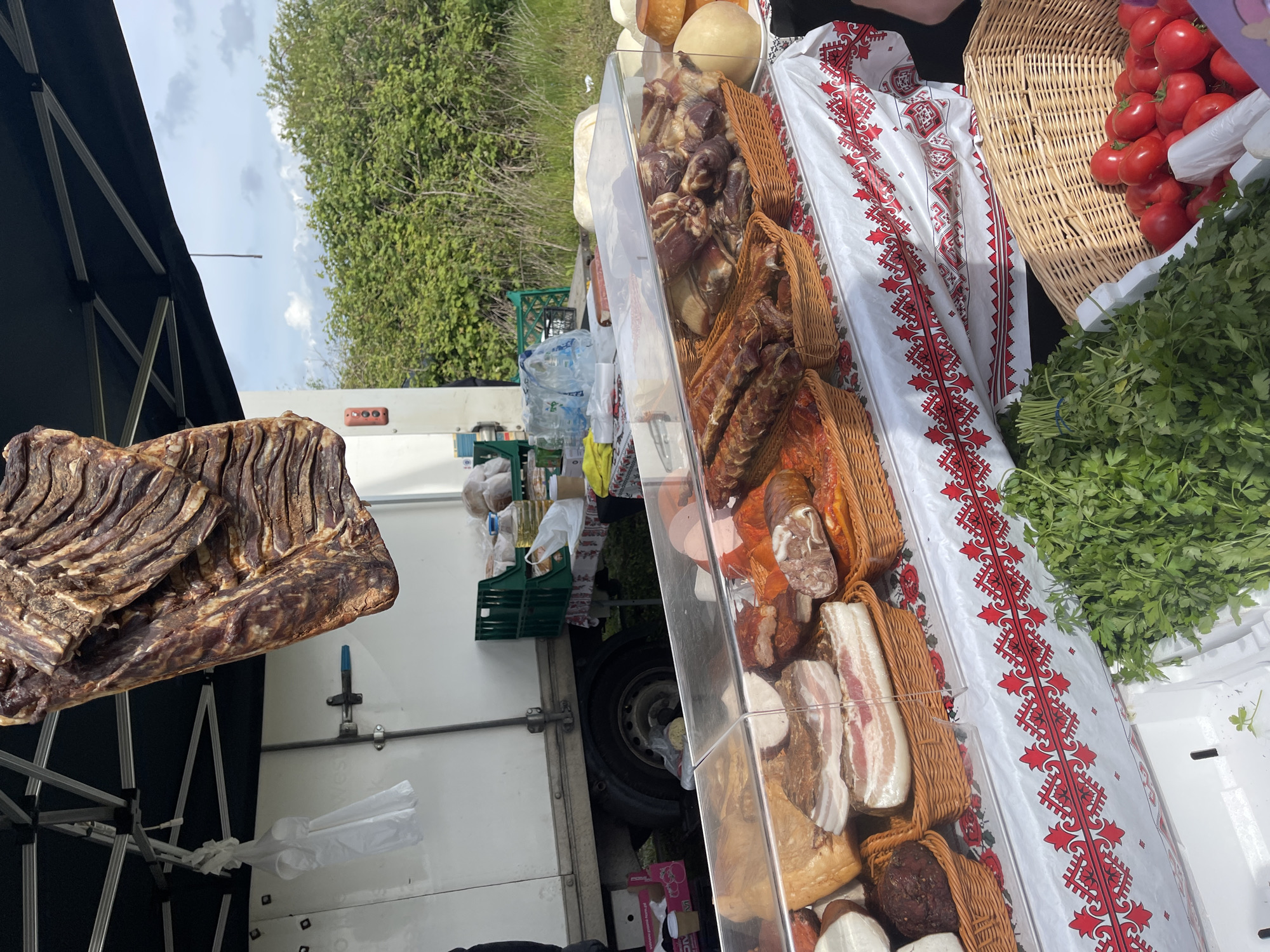 The image shows a vendor stall, likely at a market. In the foreground, a large piece of dried, cured meat hangs from the stall's frame. Behind it is a white truck with what appear to be various food items displayed in a clear case. Baskets of cured meats and cheeses are visible within the case. A white cloth with a red embroidered pattern is next to the clear case. Baskets of tomatoes and bunches of parsley are also visible. The background includes green trees and a cloudy sky. The location is unknown.