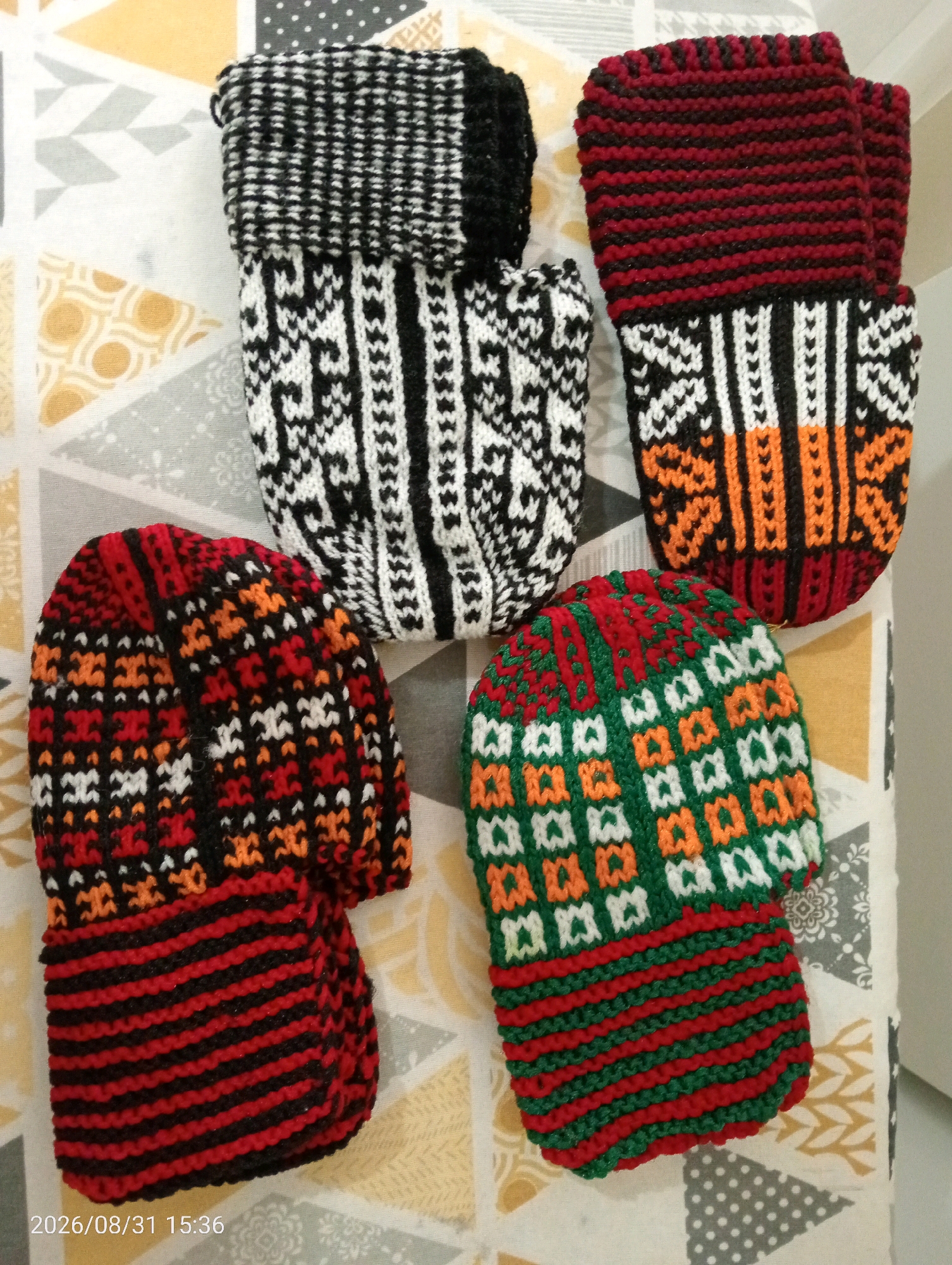 This is a close-up, top-down photograph of five knitted items, likely socks or slippers, arranged on a patterned fabric surface. The items display intricate, traditional knitting patterns in various color combinations: black and white, red and black, and green, white, and orange. The patterned fabric beneath them features geometric shapes in shades of yellow, grey, and white. A timestamp in the lower left corner reads "2026/08/31 15:36". The image is cropped to focus on the knitted items and does not show any environmental context beyond the immediate surface they are placed on. The photograph was taken indoors, as indicated by the lighting and the presence of what appears to be a wall or furniture in the upper right background. No people are visible in the image. The scene is static, with the knitted items simply arranged. There is no identifiable connection to the city of Silifke, Turkey, within the visual information provided.