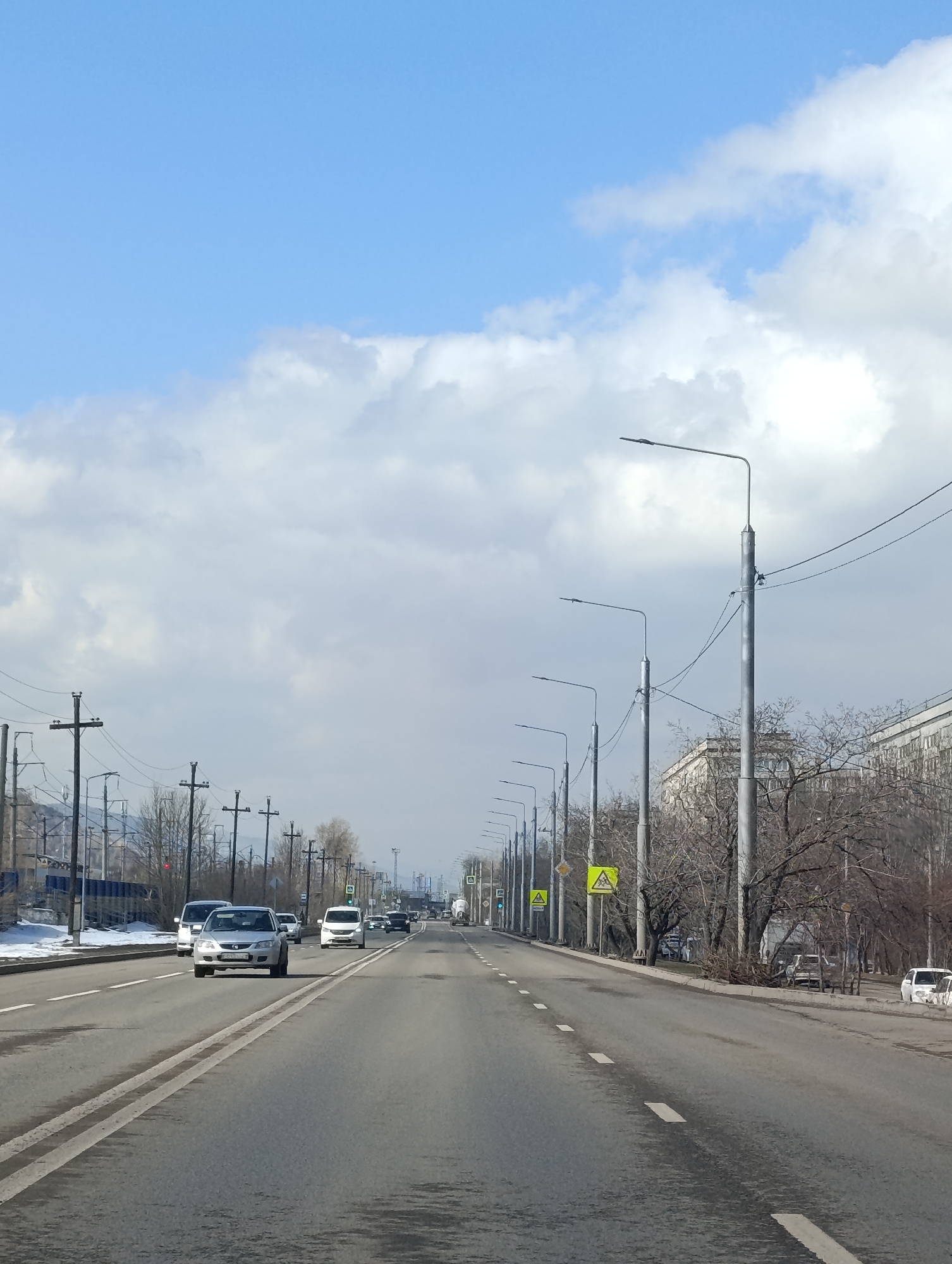 The image depicts a straight, multi-lane road running through an urban environment in Krasnoyarsk, Russia. The road surface is gray with painted white lane markings. Several vehicles are visible on the road, including a silver sedan and a white van. On either side of the road are sidewalks, bare trees, and buildings. Tall metal utility poles and streetlights line the road. The sky is light blue with scattered white clouds. Weather appears to be overcast with the sun still peeking through.