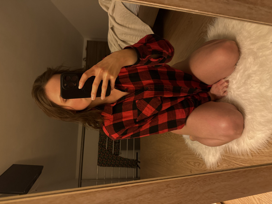 In Cava de' Tirreni, Italy, a person is observed kneeling on a white fluffy rug placed on a light wooden floor, facing a mirror to capture a selfie indoors. The individual wears an unbuttoned red and black plaid long-sleeved shirt; the lower body, visible from the mid-thigh area, is bare. A dark smartphone, held in the subject's right hand, obscures the face. The background, reflected in the mirror, depicts a room with a sloped ceiling. Visible elements include a bed with a light-colored blanket, a dark wooden dresser, a wall-mounted flat-screen television, and a patterned garment draped over a metal railing. The room is dimly illuminated with warm ambient light.