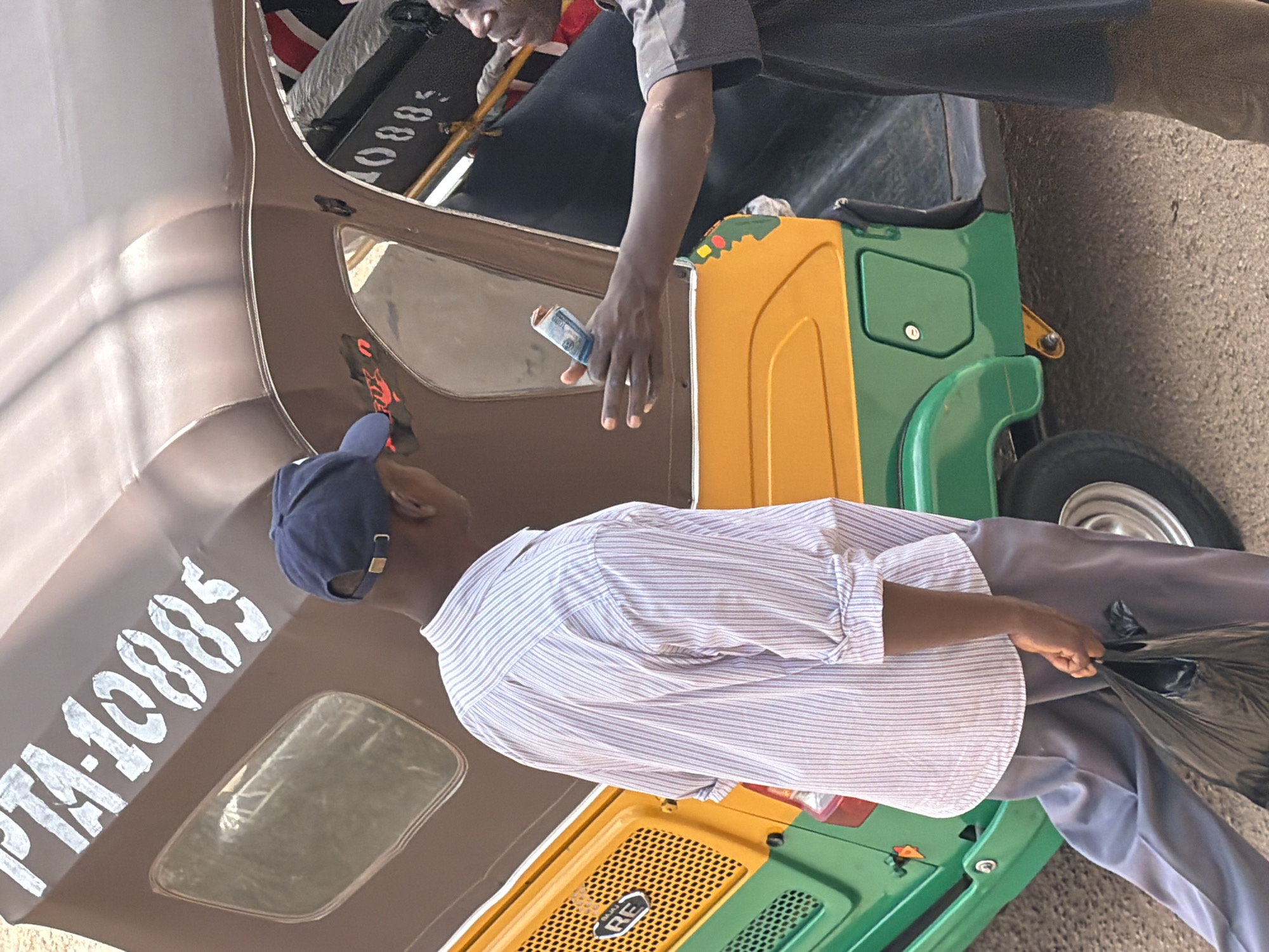 The image shows two men near a three-wheeled auto-rickshaw, possibly a form of local transportation. One man, wearing a blue baseball cap and a striped long-sleeved shirt, is facing away from the camera. He appears to be holding a black plastic bag. The other man, wearing a grey shirt, is leaning forward and holding a rolled-up banknote in his hand. The auto-rickshaw is brightly colored with yellow and green panels. White lettering on the brown part of the vehicle reads "PTA-10085". The scene appears to be outdoors, with a paved surface visible. The lighting suggests it is daytime. The overall impression is of a transaction or exchange taking place.