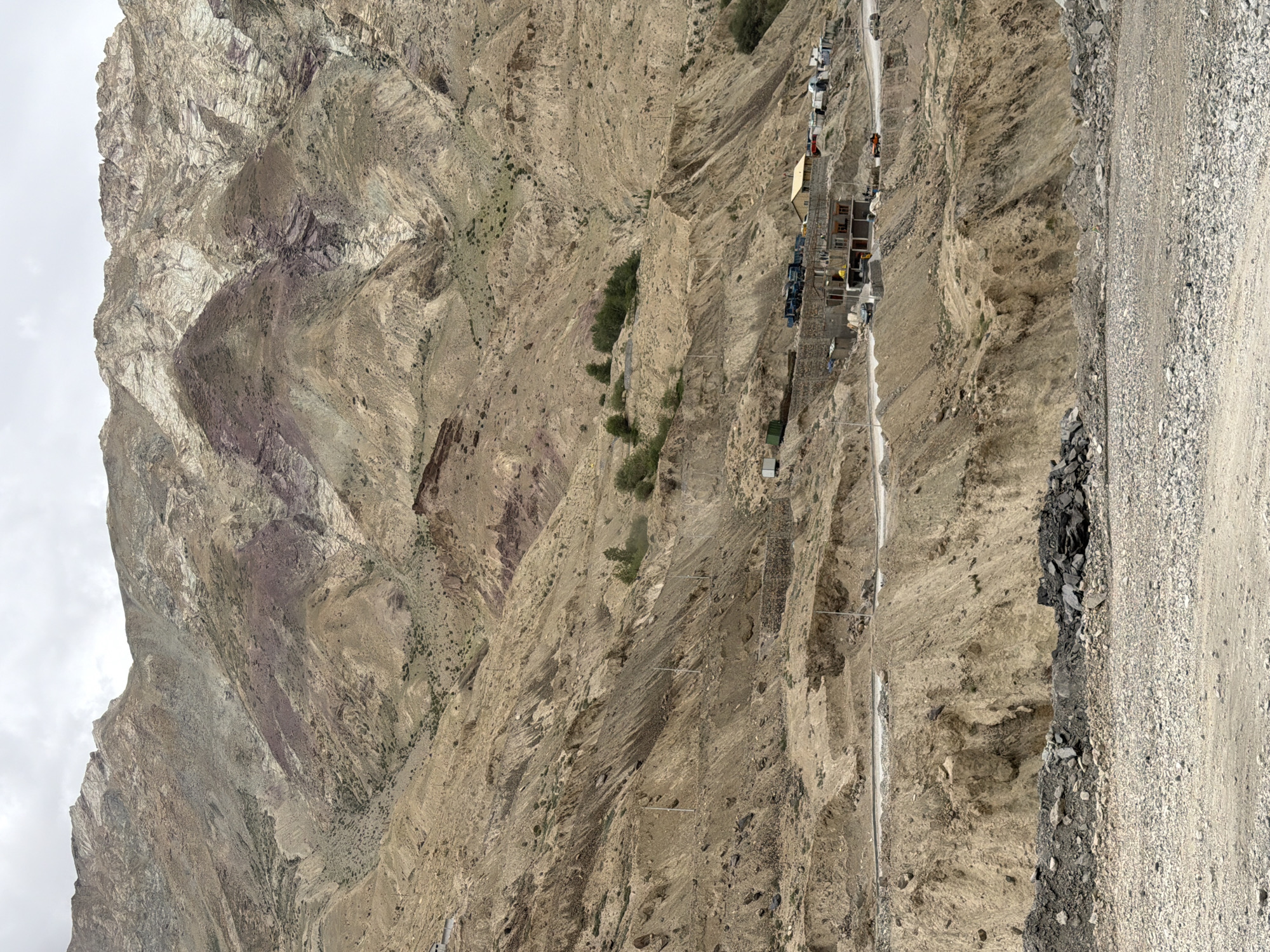 A wide, arid mountain landscape dominates the scene. The mountains are characterized by their rugged, eroded slopes, displaying layers of tan, brown, and reddish-purple rock. Sparse green vegetation clings to some of the lower slopes. A road winds through the mid-ground, carved into the mountainside, with evidence of construction activity. Several buildings, some appearing to be under construction and others more established, are visible along the road. Various vehicles, including trucks, cars, and possibly construction equipment, are parked or in motion near the buildings. The sky is overcast, suggesting a cloudy day. The foreground is covered in loose gravel and rocks.