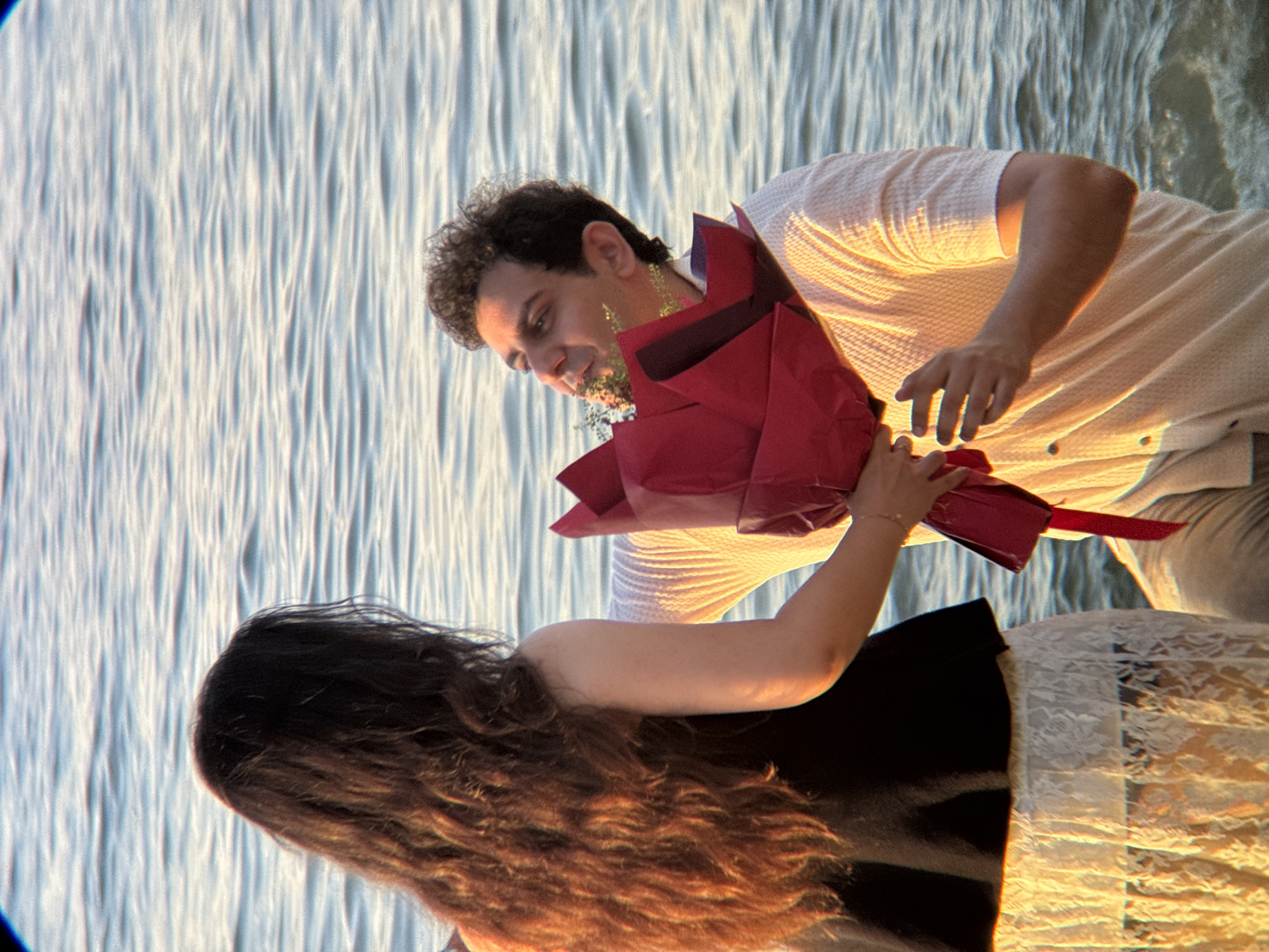 A man kneels in the shallow water of a sea, holding a large bouquet of red flowers towards a woman. The man has curly dark hair and is wearing a light-colored, textured shirt. The woman, seen from behind, has long, dark, wavy hair and is wearing a black top and a cream-colored lace skirt. The water is a light blue with gentle ripples, suggesting a calm sea. The lighting indicates it may be during sunset, casting a warm glow on the scene. The location appears to be a beach or coastline in Nha Trang, Vietnam, a popular tourist destination known for its beautiful beaches and clear waters.