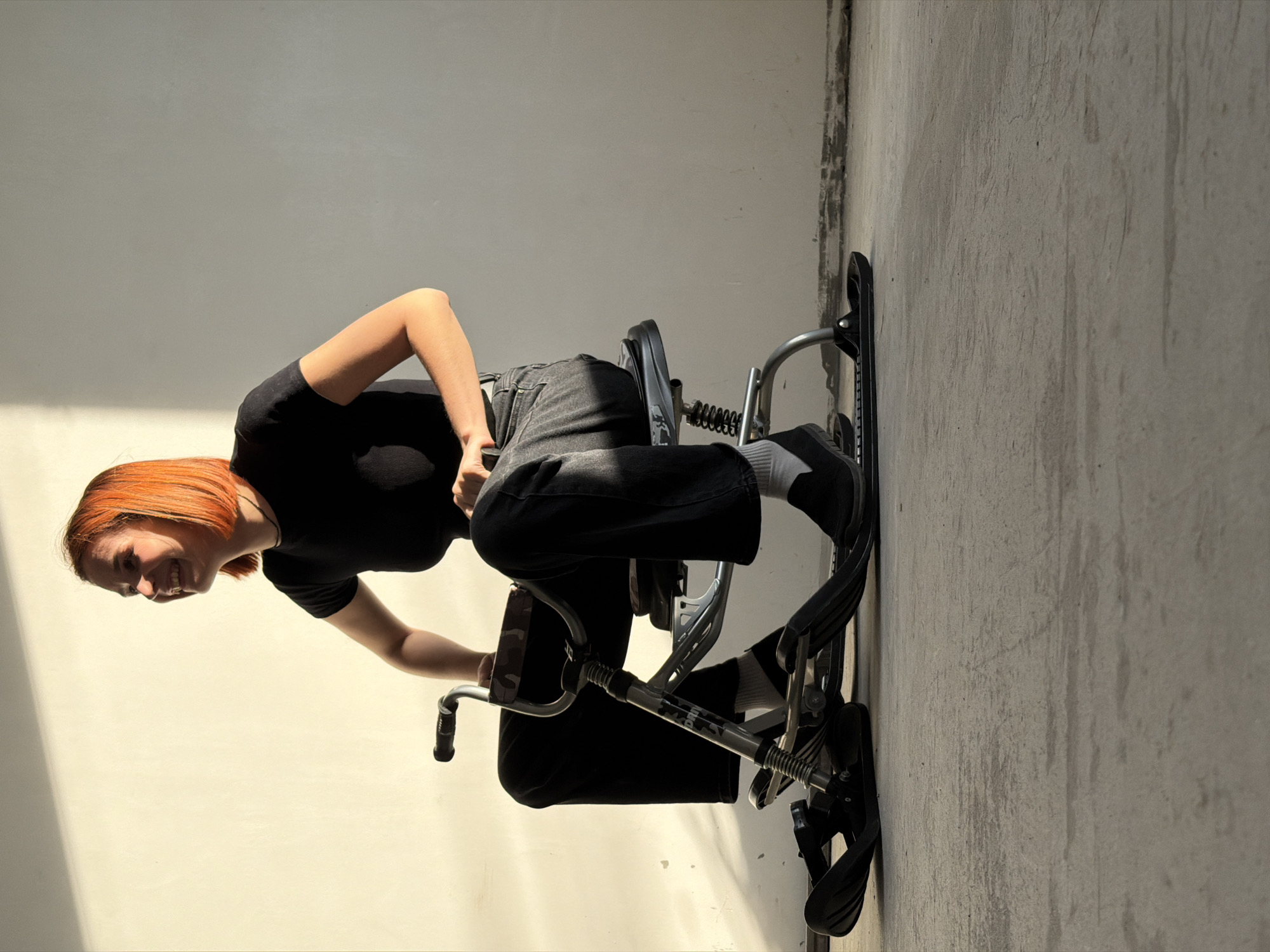 A woman with red hair is sitting on a small wheeled vehicle indoors. She is wearing a black t-shirt, dark jeans and white socks, and is smiling. The vehicle has a grey metal frame with a seat and handlebars. The background consists of two walls: a light-colored one and a darker, textured one. The picture was taken in the city of Chaikovsky, Russia.