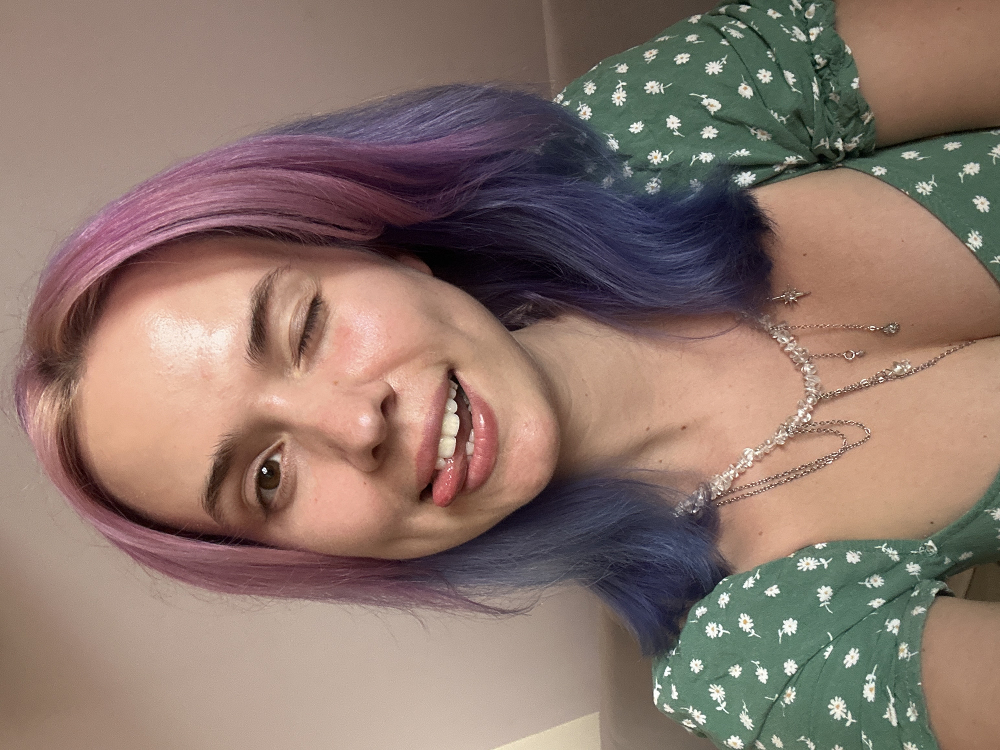 A young adult woman is captured in a close-up portrait, angled towards the viewer. She winks one eye and sticks out her tongue in a playful expression. Her hair is dyed in a vibrant two-tone style, transitioning from pink to blue. She wears a green dress with small white floral patterns. A delicate silver necklace adorns her neck. The background is a plain, light-colored wall. The setting's location in the city of Gijón/Xixón, Spain is not identifiable from the visual information provided.