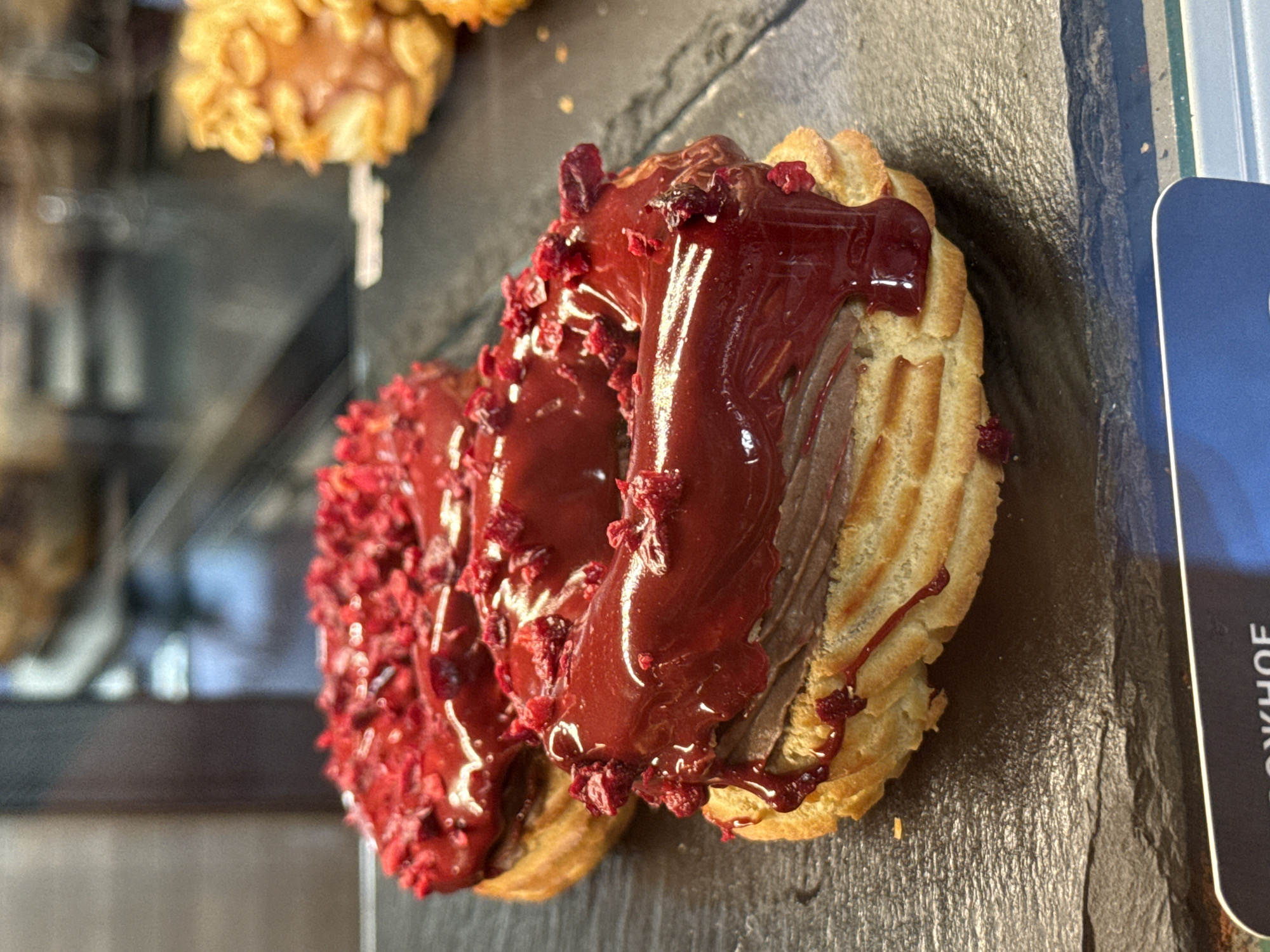 This media file features a close-up, slightly angled shot of two éclairs, presented on a dark, textured surface, likely a slate platter. The éclairs are ring-shaped and appear to be topped with a rich, dark red glaze, possibly raspberry or cherry, with visible dried fruit pieces sprinkled on top. The glaze is slightly drippy, suggesting freshness. Between the glaze and the pastry, there is a filling that appears to be a chocolate cream. The pastry itself is golden brown and has the characteristic ridged texture of choux pastry.
In the background, slightly out of focus, are other baked goods, possibly more éclairs or donuts, with a lighter, golden-brown color and some kind of crunchy topping. The setting appears to be a display case or counter in a bakery or cafe. To the right of the éclairs, a corner of a blue card with white text is visible. The visible text is in Cyrillic and reads "ОЖНОЕ". Given the location context of Saint Petersburg, Russia, this card is likely related to branding or product information.
The lighting is bright, highlighting the glossy texture of the glaze and the details of the pastry. There are no people visible in the frame. The overall impression is of a tempting display of sweet treats, emphasizing their texture and appealing colors. The scene suggests a daytime setting, typical of a commercial establishment selling food.