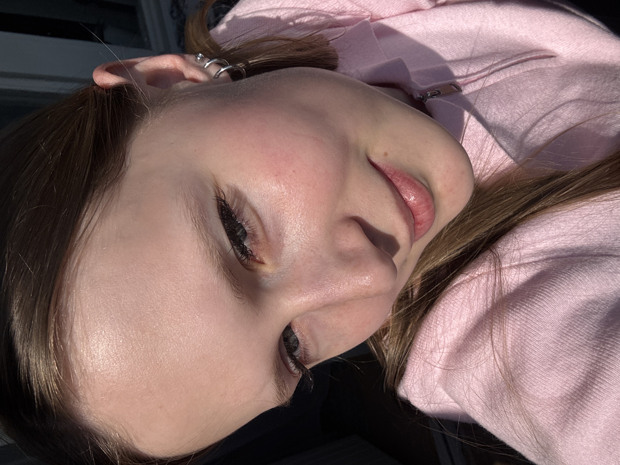 A close-up, angled portrait captures the face and upper body of a young person in Chaikovsky, Russia. The subject, likely female, has fair skin, light brown hair, and is wearing a pink hooded sweatshirt with a visible silver zipper pull. Their head is tilted, and the photo is taken from an elevated perspective, looking down slightly.
The left side of the subject's face is prominent, featuring a partially visible eye with long, dark eyelashes. Their lips are slightly parted, revealing a hint of the inner lip. The ear is adorned with multiple silver piercings, including hoops on the lobe and helix, and a stud on the upper cartilage. Strong, directional sunlight illuminates the subject, creating bright highlights on the skin, hair, and the textured fabric of the sweatshirt, while also casting distinct shadows, particularly under the nose and chin, suggesting it is daytime with direct sun exposure. The hair, a mix of light brown and auburn tones, falls around the face and on the shoulder, with some strands catching the light. The background is dark and out of focus, providing no specific location cues beyond the provided context. The person's expression appears neutral or contemplative, posing for the camera. There is no visible text in the image.