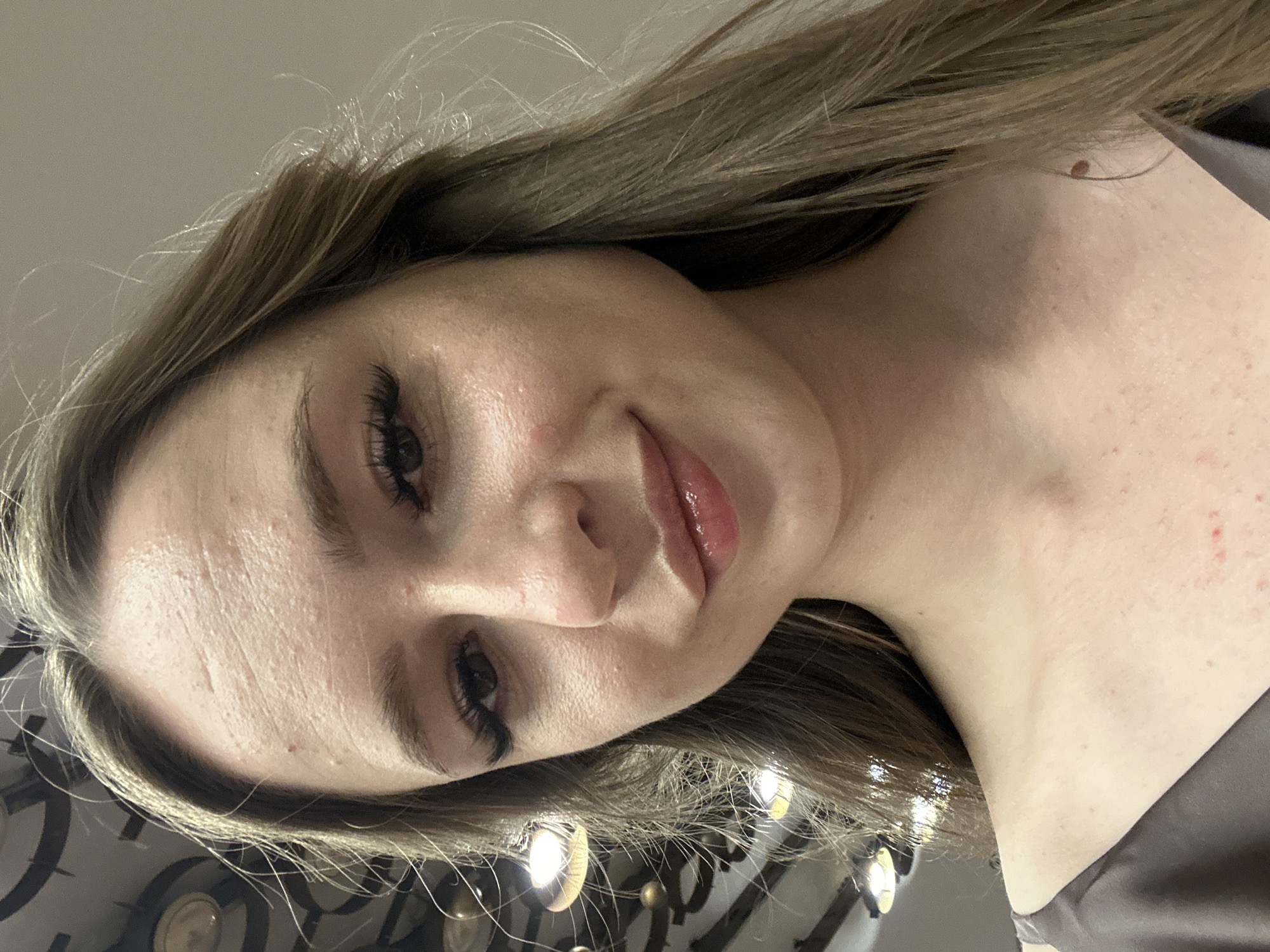 An upward-angle, close-up selfie captures a young adult woman with long, wavy blonde hair. She is wearing a dark grey, satin-like top. Her face is the primary focus, with visible makeup including dramatic false eyelashes and lip gloss. Her skin appears slightly oily and shows some texture and redness on her forehead, cheeks, and chest. The background is blurred and shows what appear to be ceiling lights and some structural elements, possibly suggesting an indoor setting like a retail store or event space. The lighting is bright and appears to be coming from overhead.