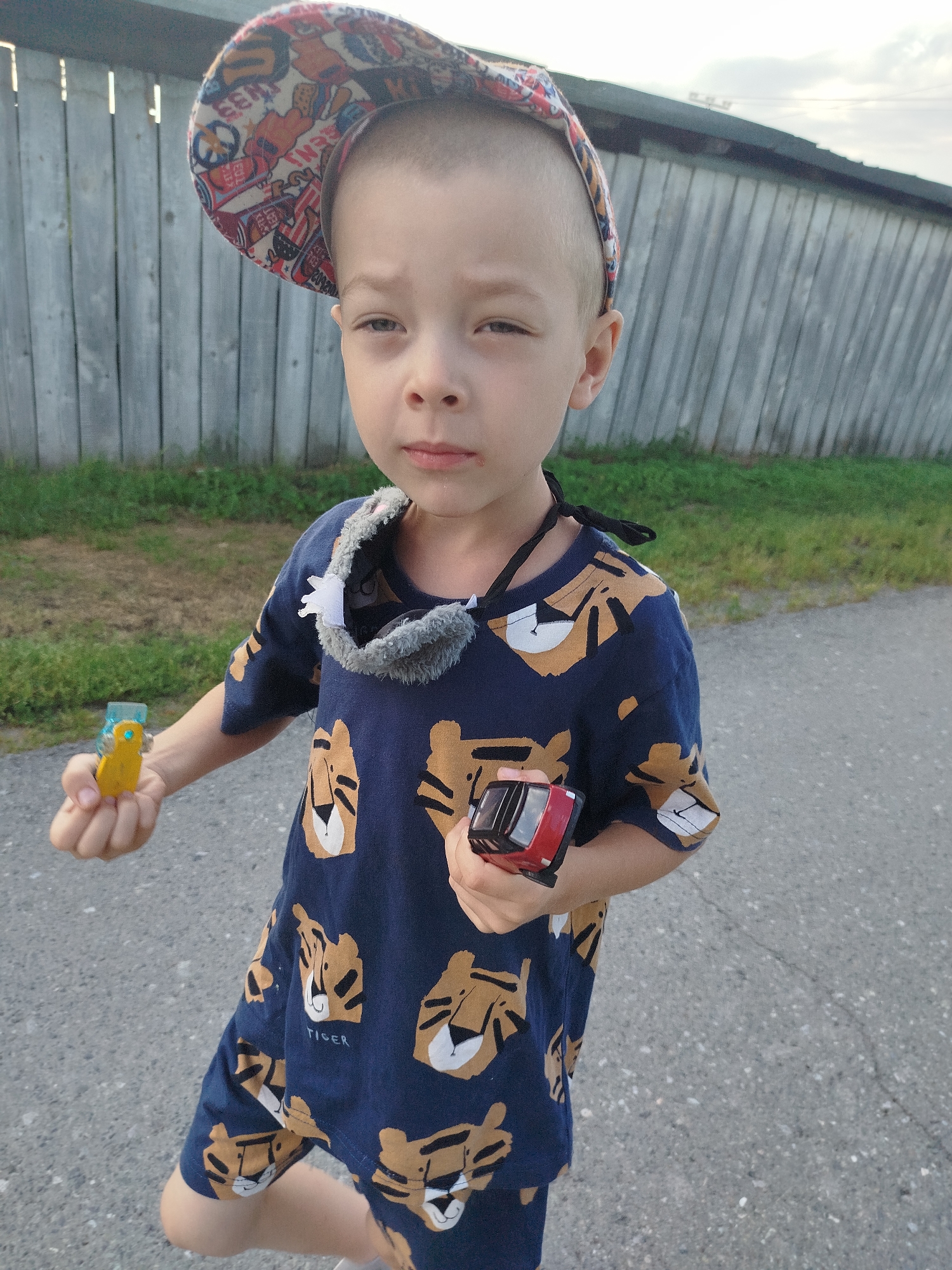 A young boy with a shaved head wears a multi-colored baseball cap and a dark blue t-shirt with a repeating pattern of tiger faces. He has a gray, fuzzy scarf or bandana around his neck. In his left hand, he holds a small yellow toy car, and in his right hand, he holds a small red toy car. The boy is standing on an asphalt surface, possibly a sidewalk or road, with a wooden fence in the background. Green grass is visible between the asphalt and the fence. The lighting suggests it is daytime, likely overcast or late afternoon. The scene appears to be outdoors, possibly in a residential area of Zavodoukovsk, Russia, given the presence of a fence and grassy area.