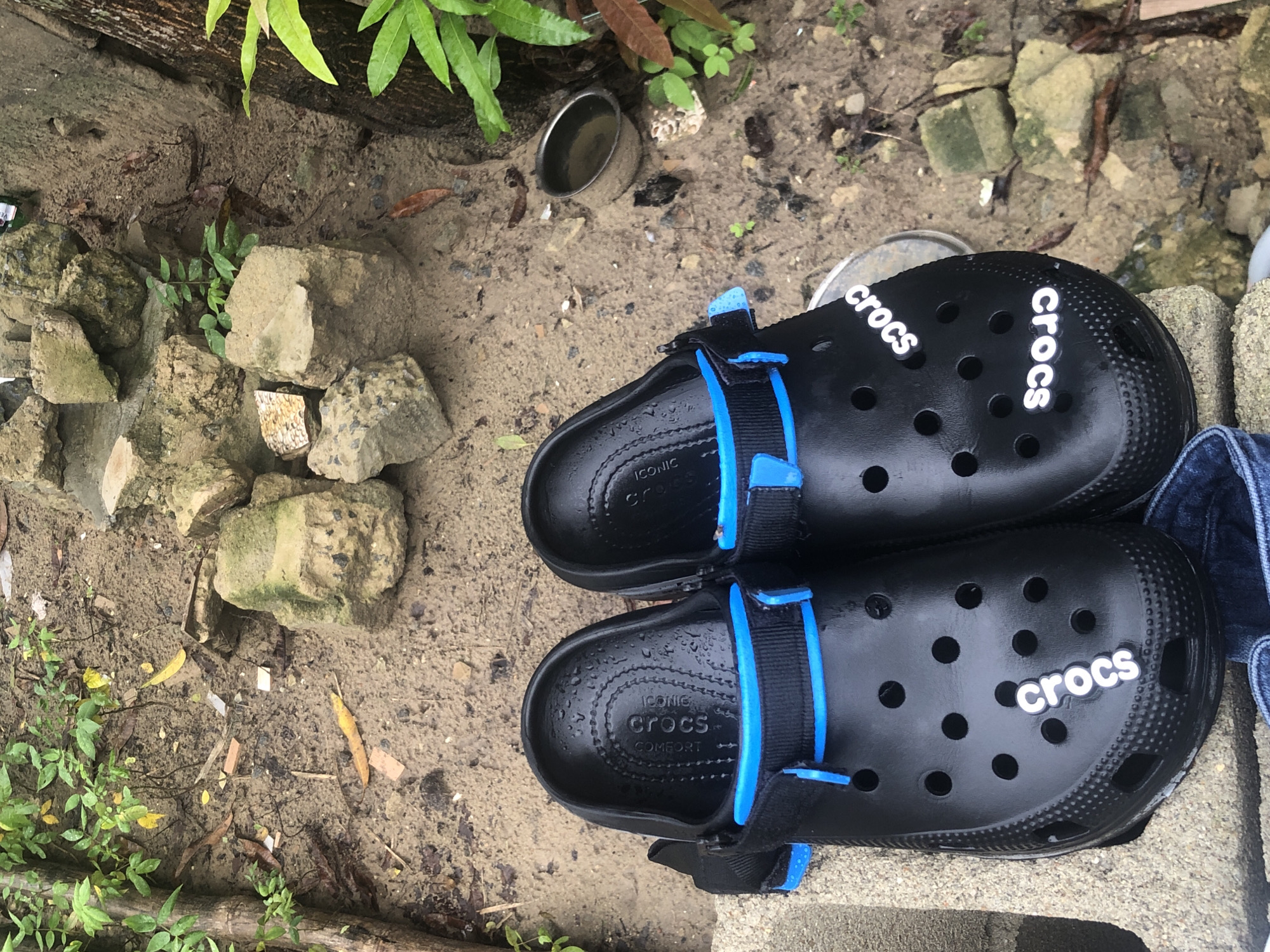 A pair of black Crocs with blue straps sits on a concrete block in an outdoor setting. The Crocs are placed next to each other. The surrounding environment consists of dirt, rocks, and some greenery. A metal cup is visible in the background. Jeans are visible to the right of the Crocs. The scene appears to be in Ado, Nigeria.