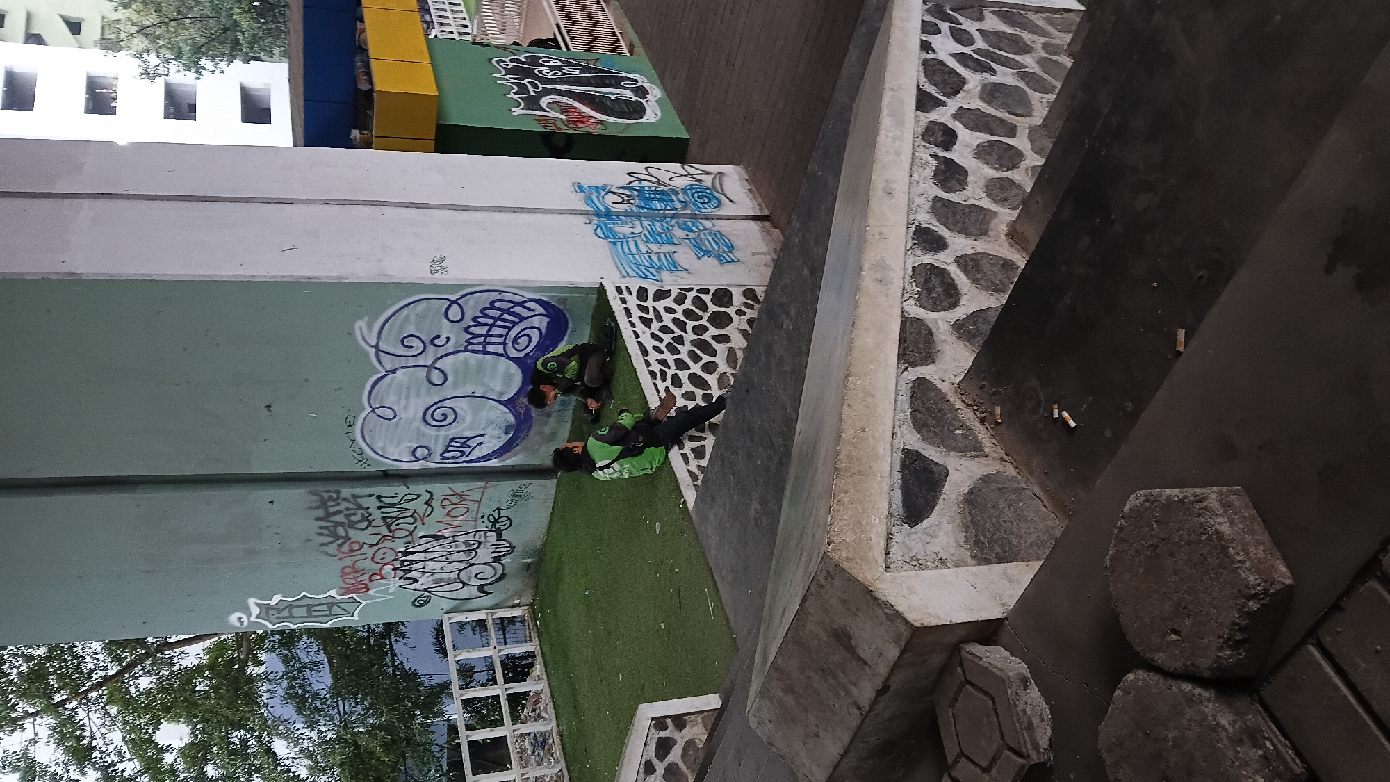 Two men in green and black uniforms are sitting on a raised grassy area under a concrete structure. The concrete structure is covered in graffiti, with various tags and drawings visible. The men are facing each other and appear to be in conversation. One man is wearing a green jacket with black sleeves and a black bag on his back. The other man is wearing a green jacket and dark pants. The grassy area is bordered by a stone wall with a patterned design. In the foreground, there are concrete steps with some discarded cigarette butts on them. The background shows a multi-story building and trees. The scene appears to be an urban outdoor space, possibly a public area or underpass, in Bandung City, Indonesia.