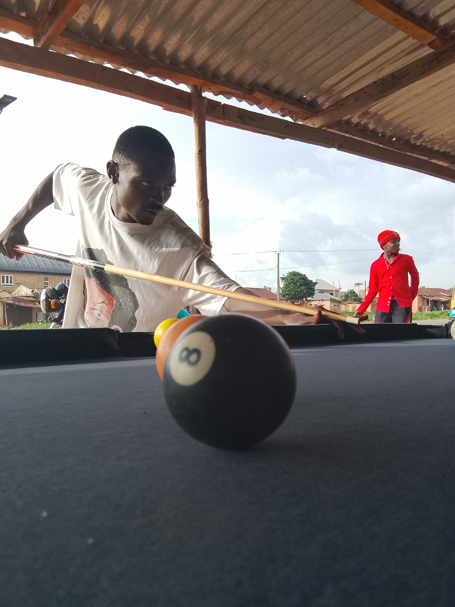 A man in a white t-shirt is aiming at pool balls with a pool cue on a pool table. Another man in a red shirt and head covering stands nearby, watching. The scene is set under a corrugated metal roof. Outside the structure, there's a glimpse of a town with houses, trees, and utility poles. The location appears to be in a city.